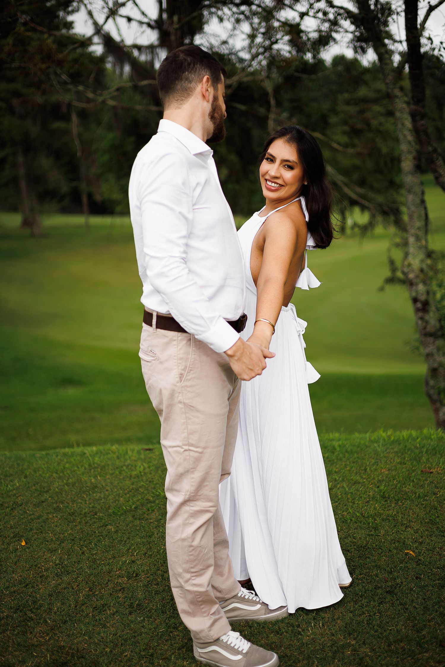 A smiling couple in white outfits holding hands on a lush green lawn surrounded by trees.