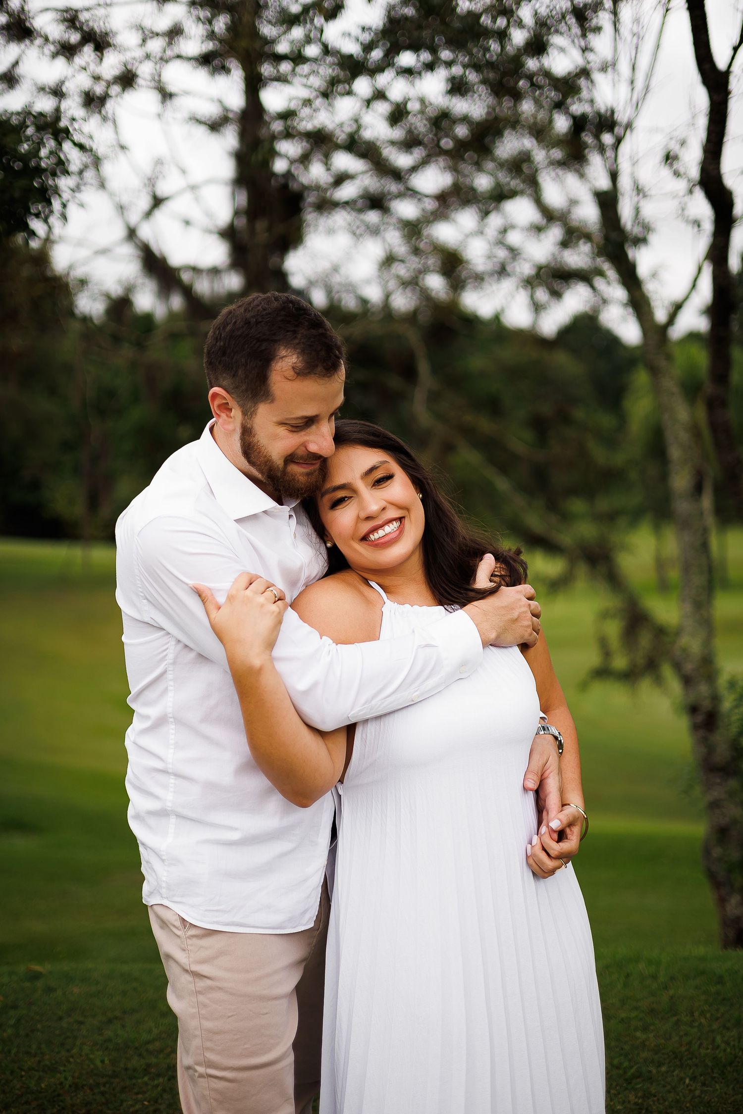 Couple in white outfits embracing outdoors, smiling woman in white dress hugged by man in white shirt on green lawn.