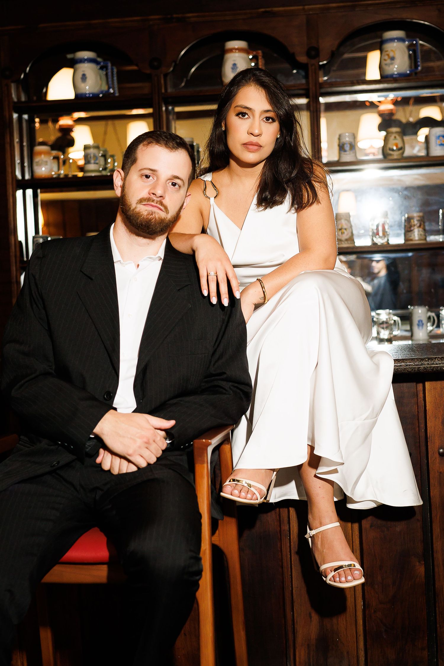 A man in a black suit and woman in white jumpsuit pose elegantly at a vintage bar setting.
