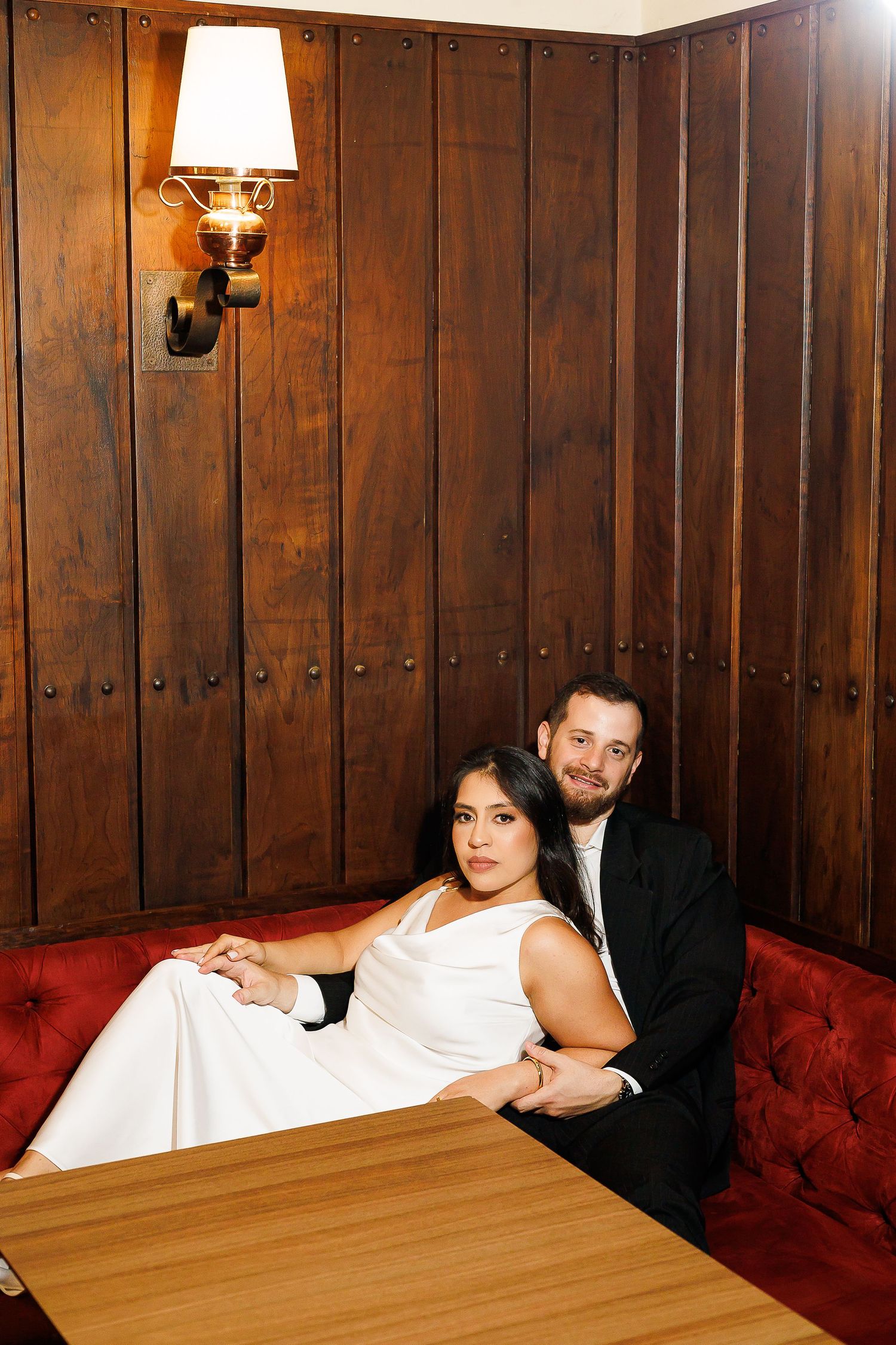 Bride and groom sitting in a red velvet booth at a wood-paneled restaurant, posing for a wedding portrait.