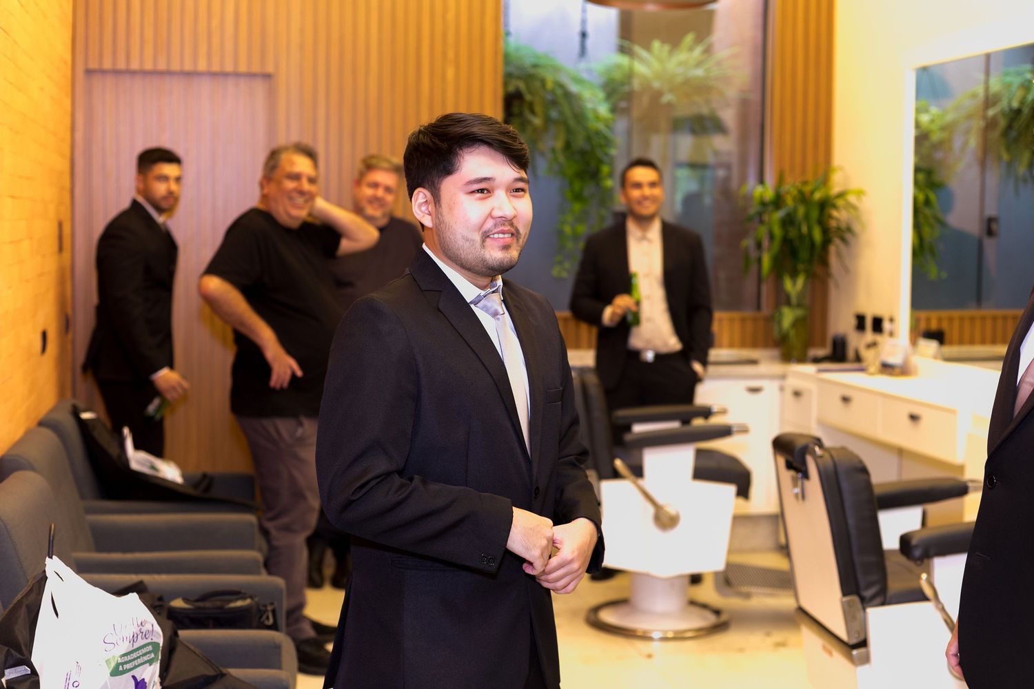 Man in black suit smiling in modern barbershop with stylish chairs and group of people in background.