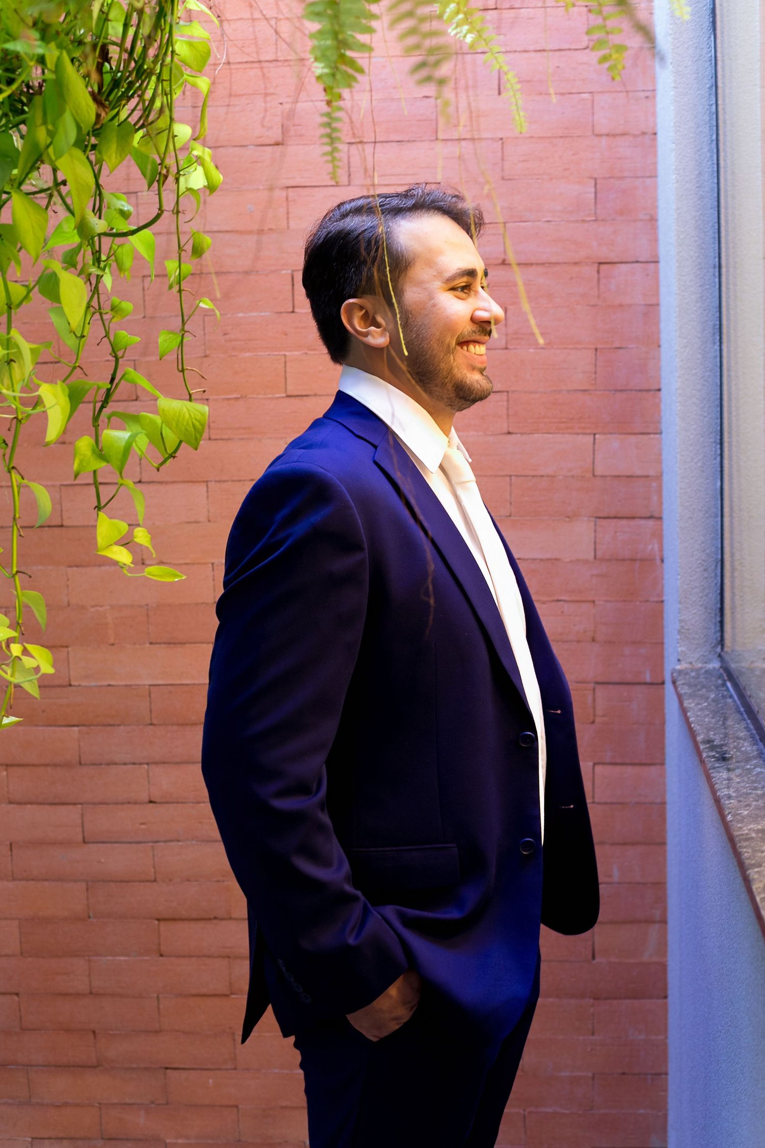 Smiling man in navy blue suit standing confidently against a red brick wall with green foliage.