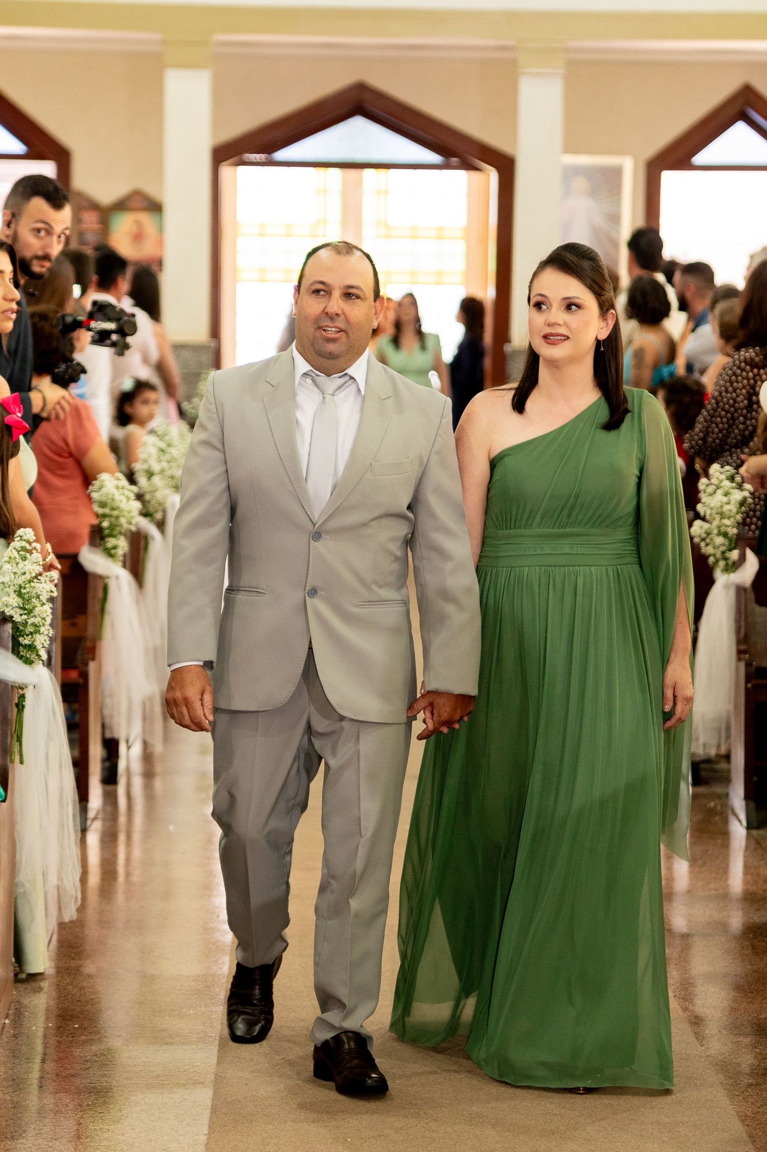 Man in gray suit and woman in green gown walk down church aisle at wedding ceremony.