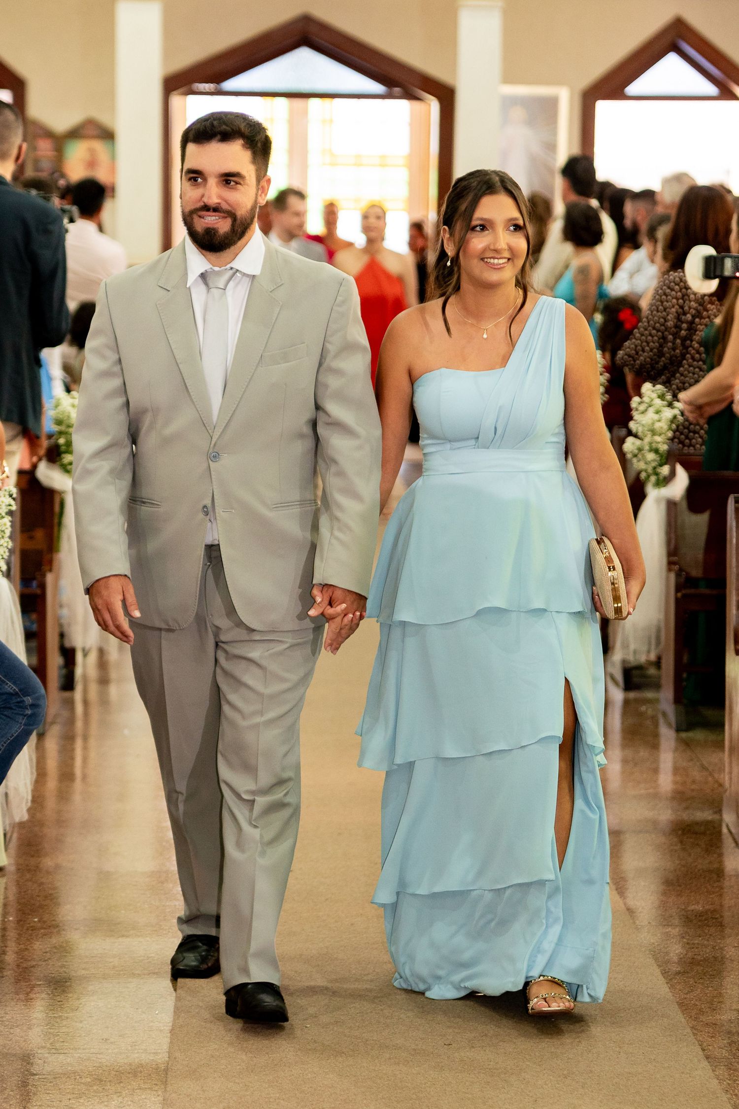 Couple walking down church aisle, man in gray suit, woman in light blue tiered one-shoulder gown at a wedding.