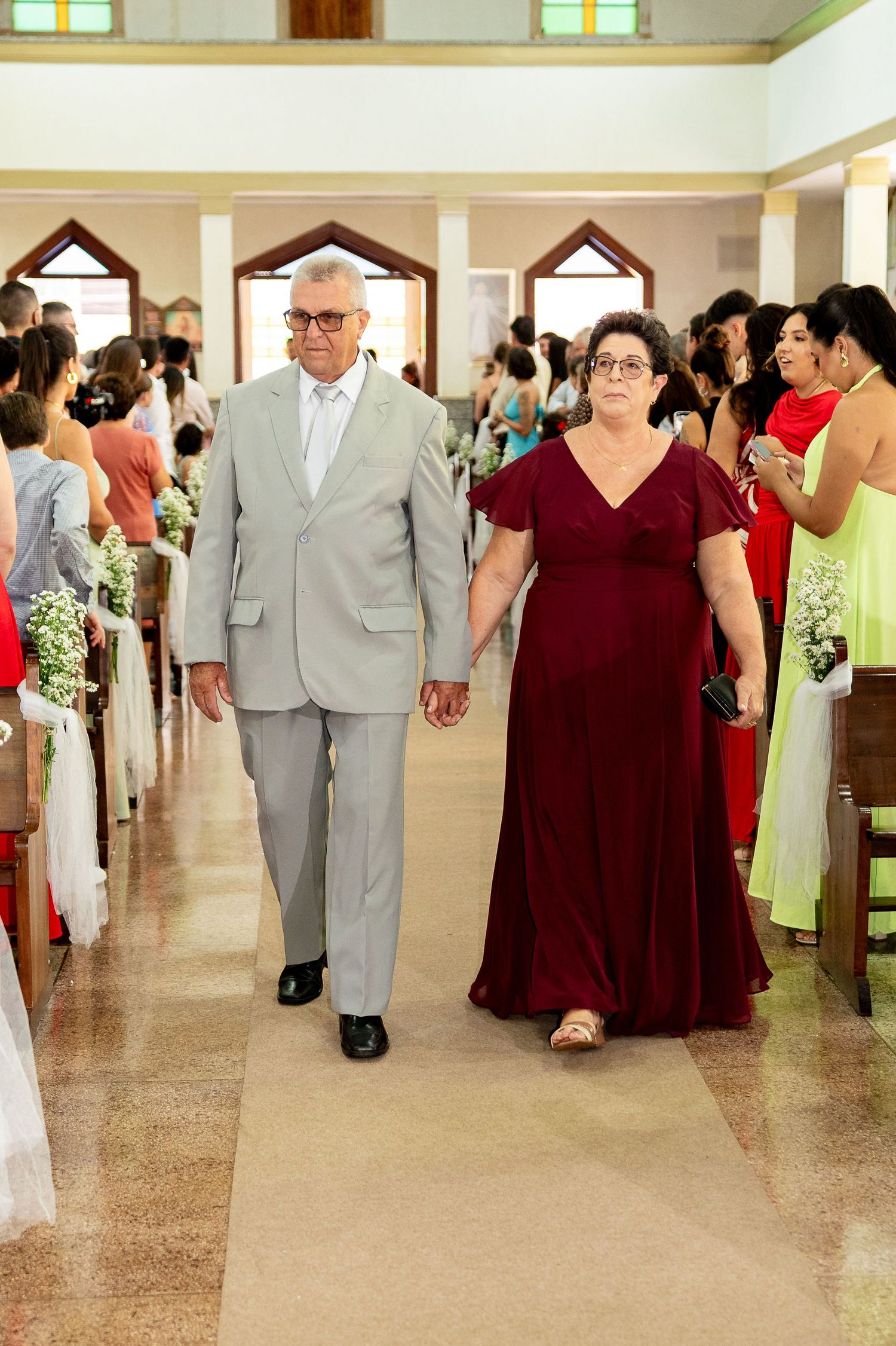 Elderly couple walking hand in hand down church aisle at wedding, man in gray suit, woman in burgundy gown.