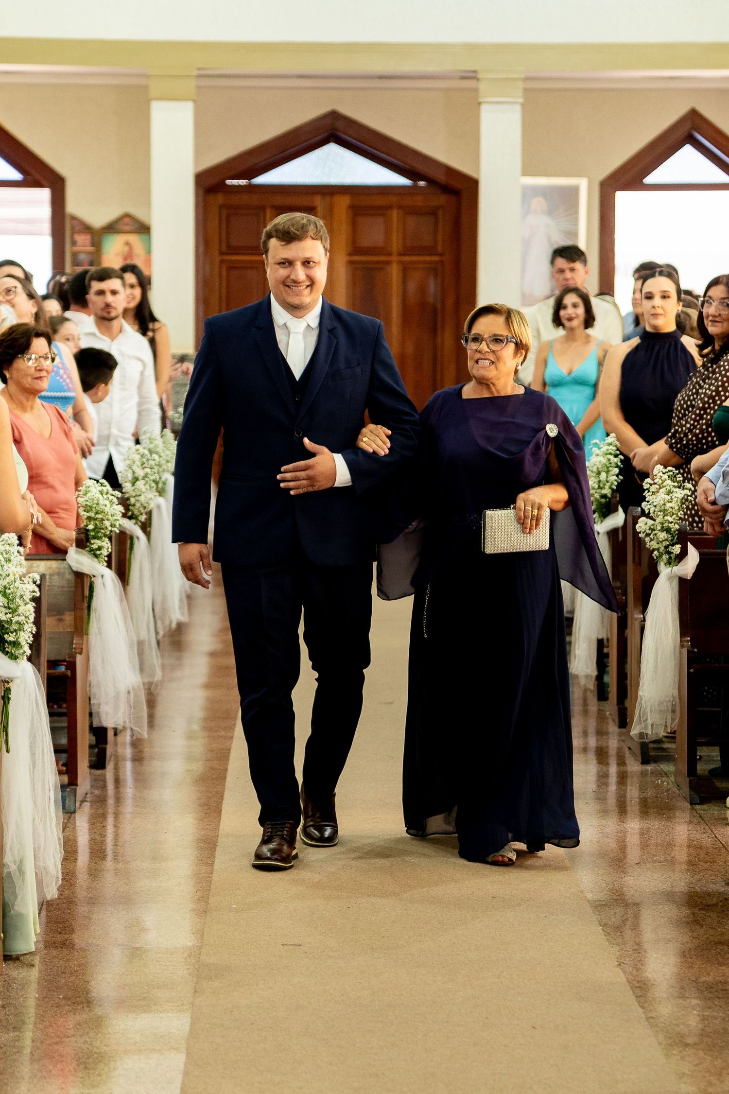 Groom in navy suit walks down church aisle with mother in purple dress during wedding ceremony.