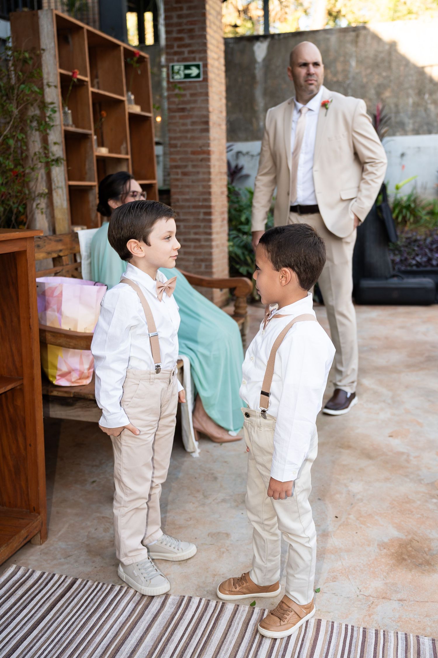 Two boys in matching suspenders and bow ties stand talking at an outdoor wedding venue with an adult man in a beige suit behind them.
