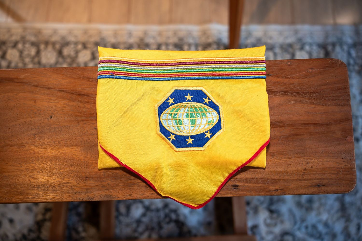 Yellow folded neckerchief with globe emblem patch and colorful striped border displayed on wooden surface.