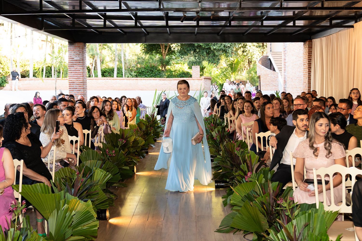 Woman in light blue gown walks down floral-lined aisle at elegant outdoor wedding ceremony with seated guests.