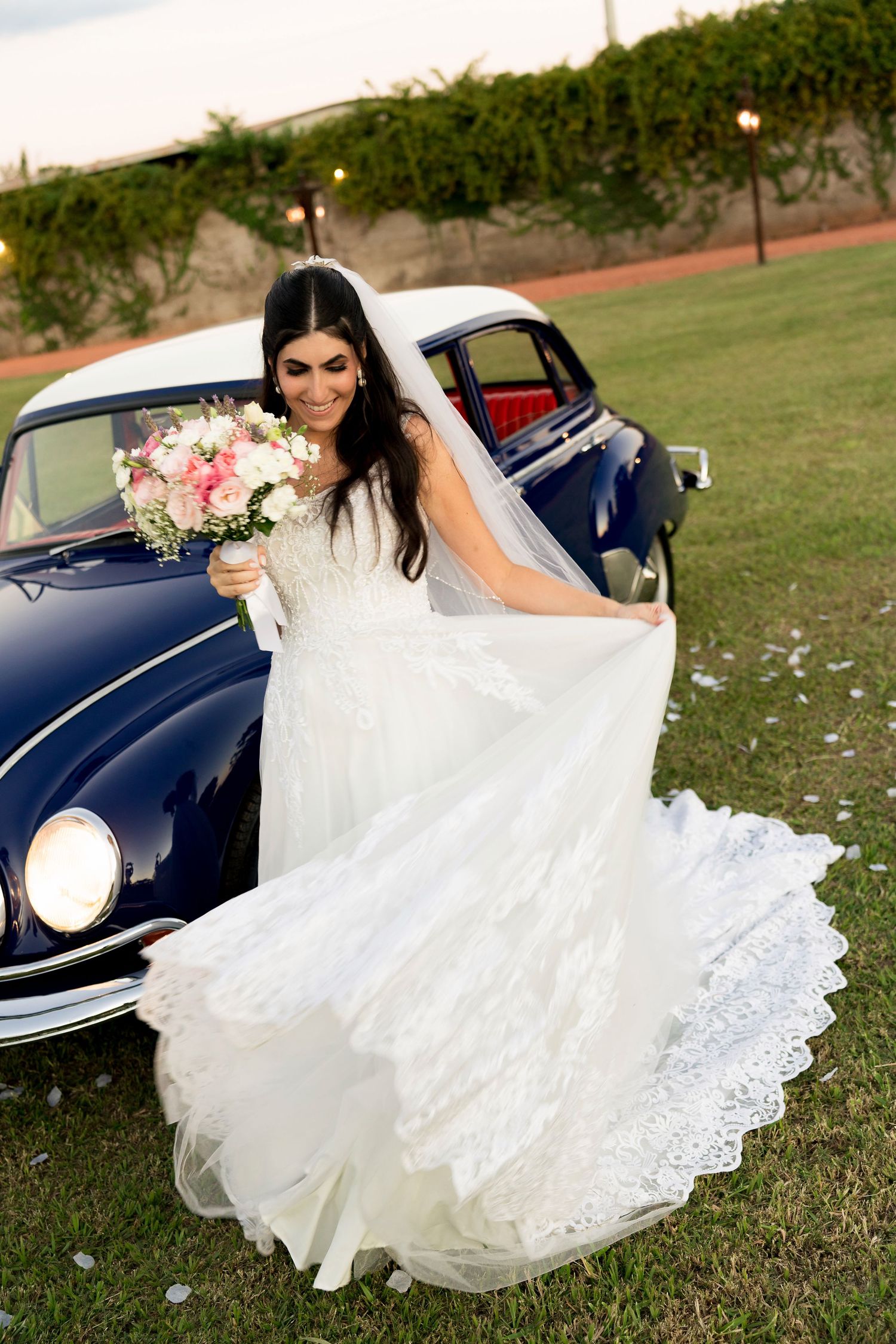 Bride in white lace gown holding pink bouquet, standing by vintage navy blue car on green lawn.