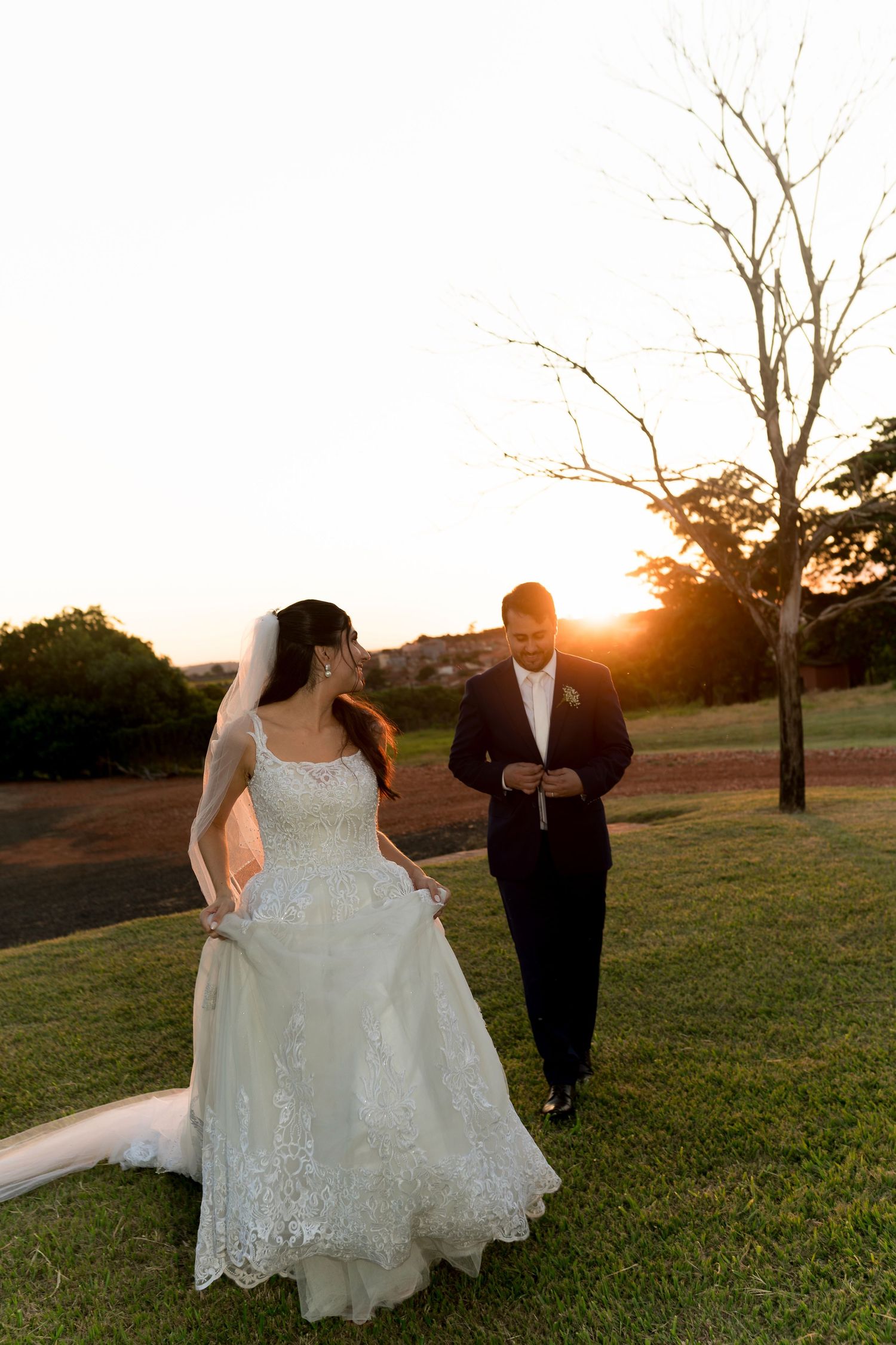 Bride in lace gown and groom in black tuxedo walk together on green lawn at golden sunset.