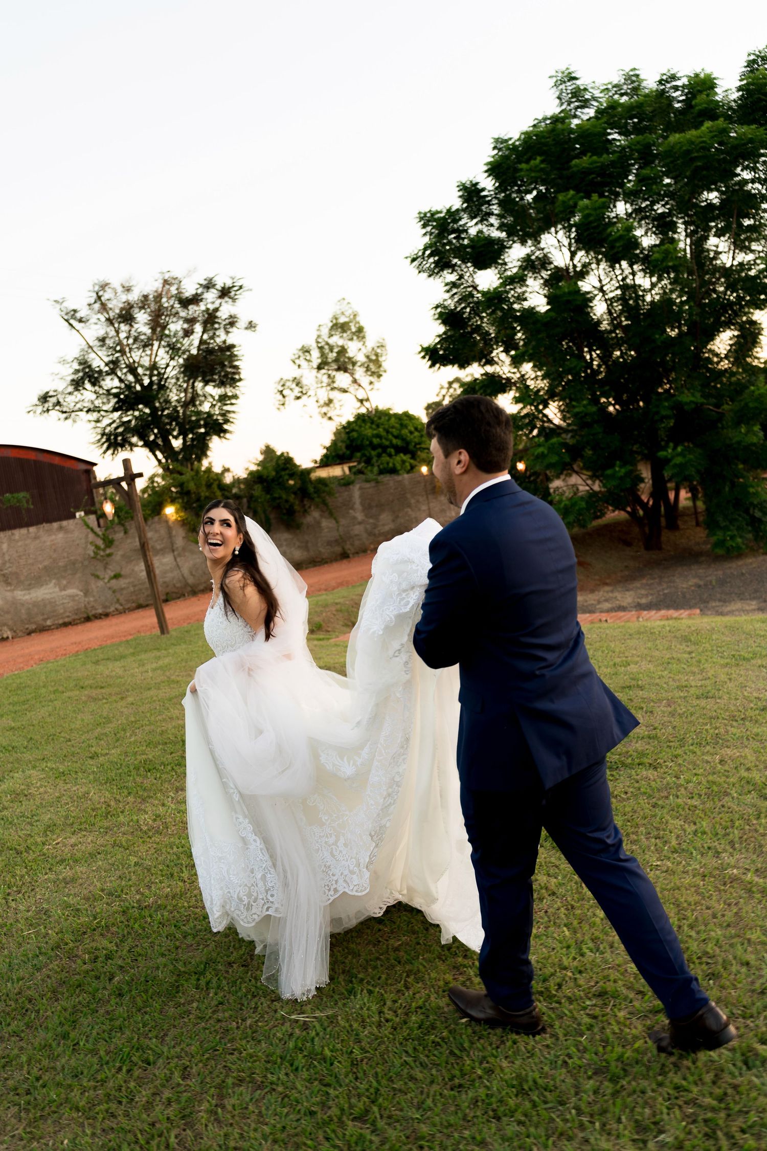 A joyful bride in a white lace gown laughs as her groom in a navy suit twirls her on a green lawn at sunset.