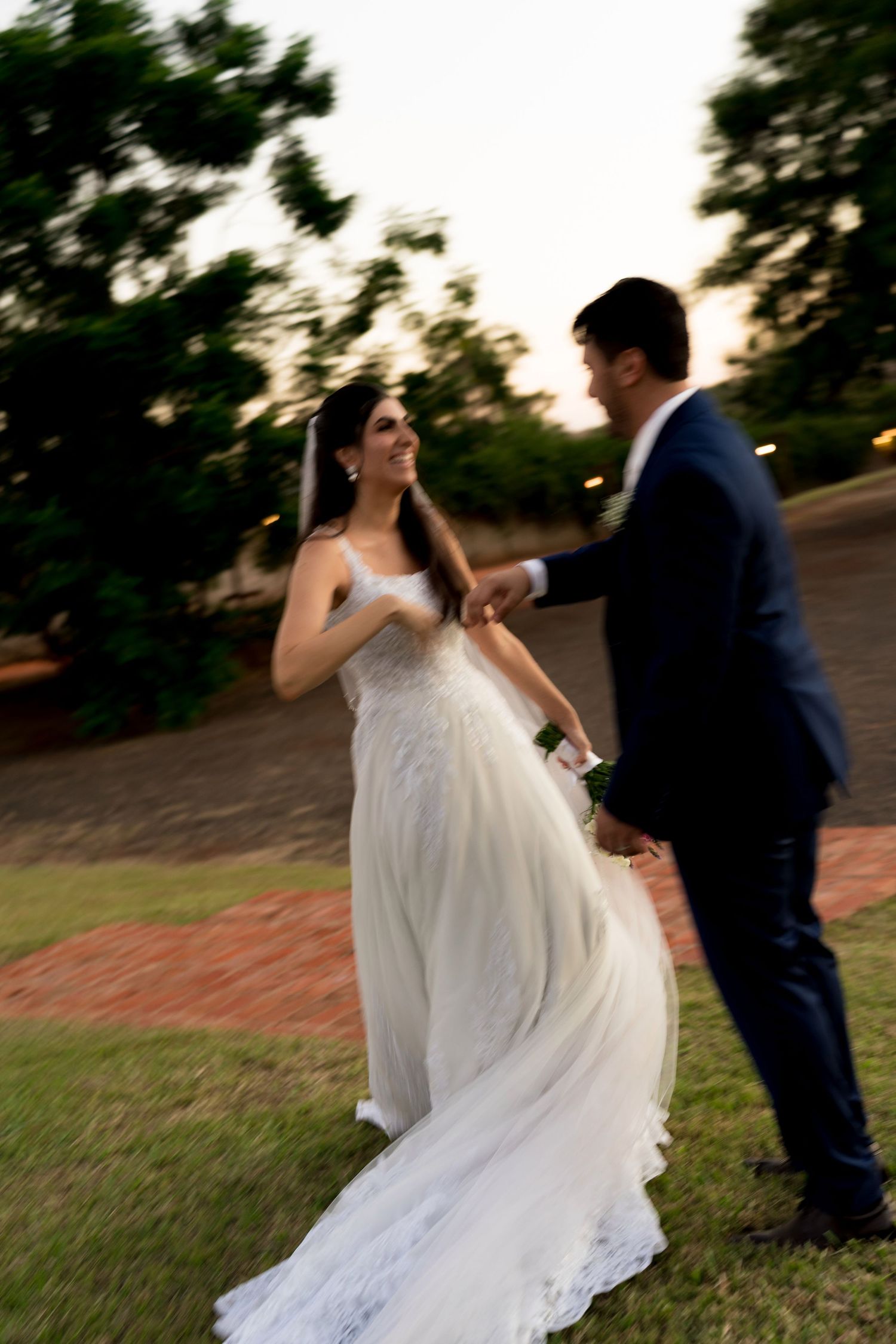 A joyful bride in a white lace gown dances with her groom in a navy suit at an outdoor wedding.