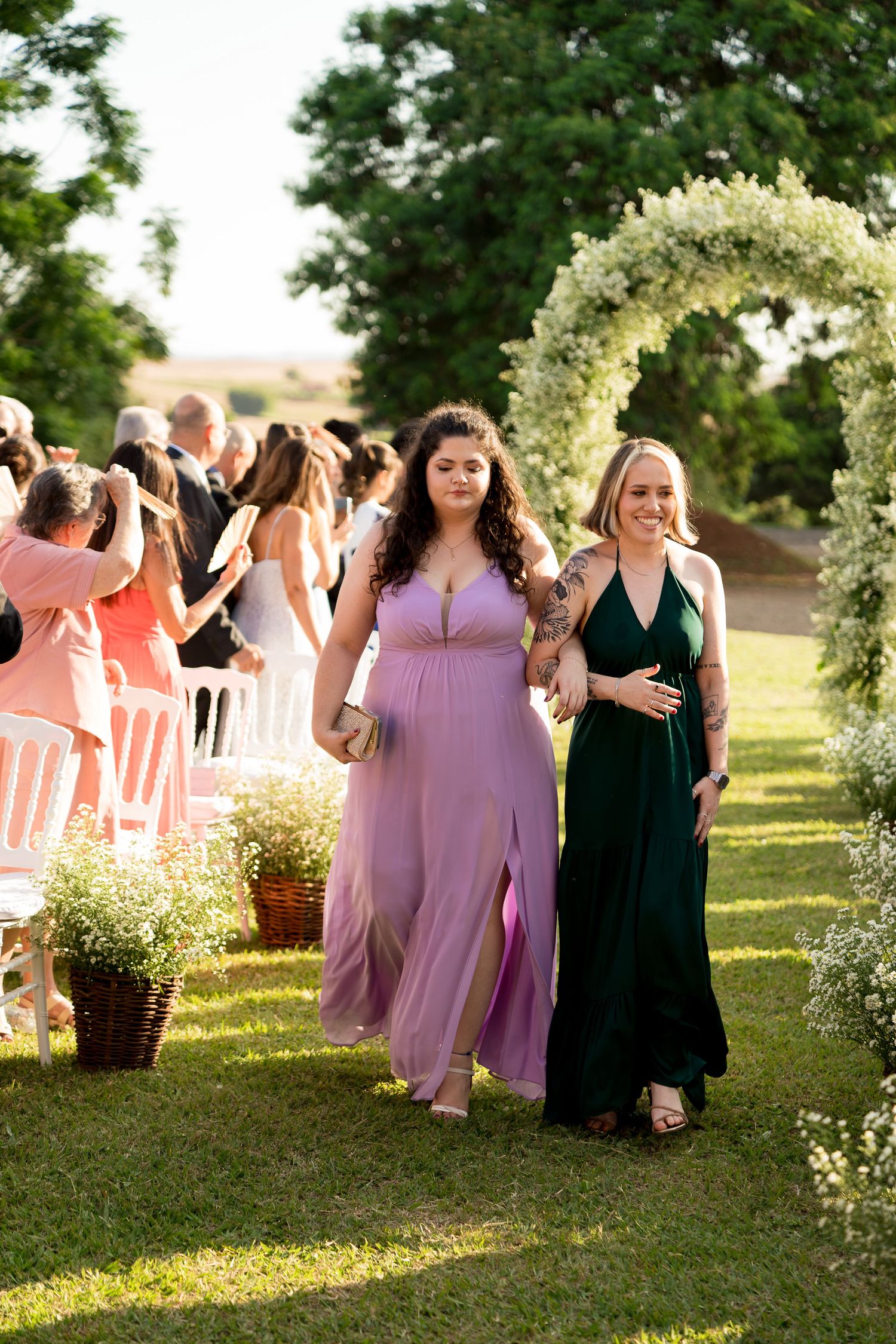 Two women in elegant gowns walk down an outdoor wedding aisle under a floral arch on a sunny day.