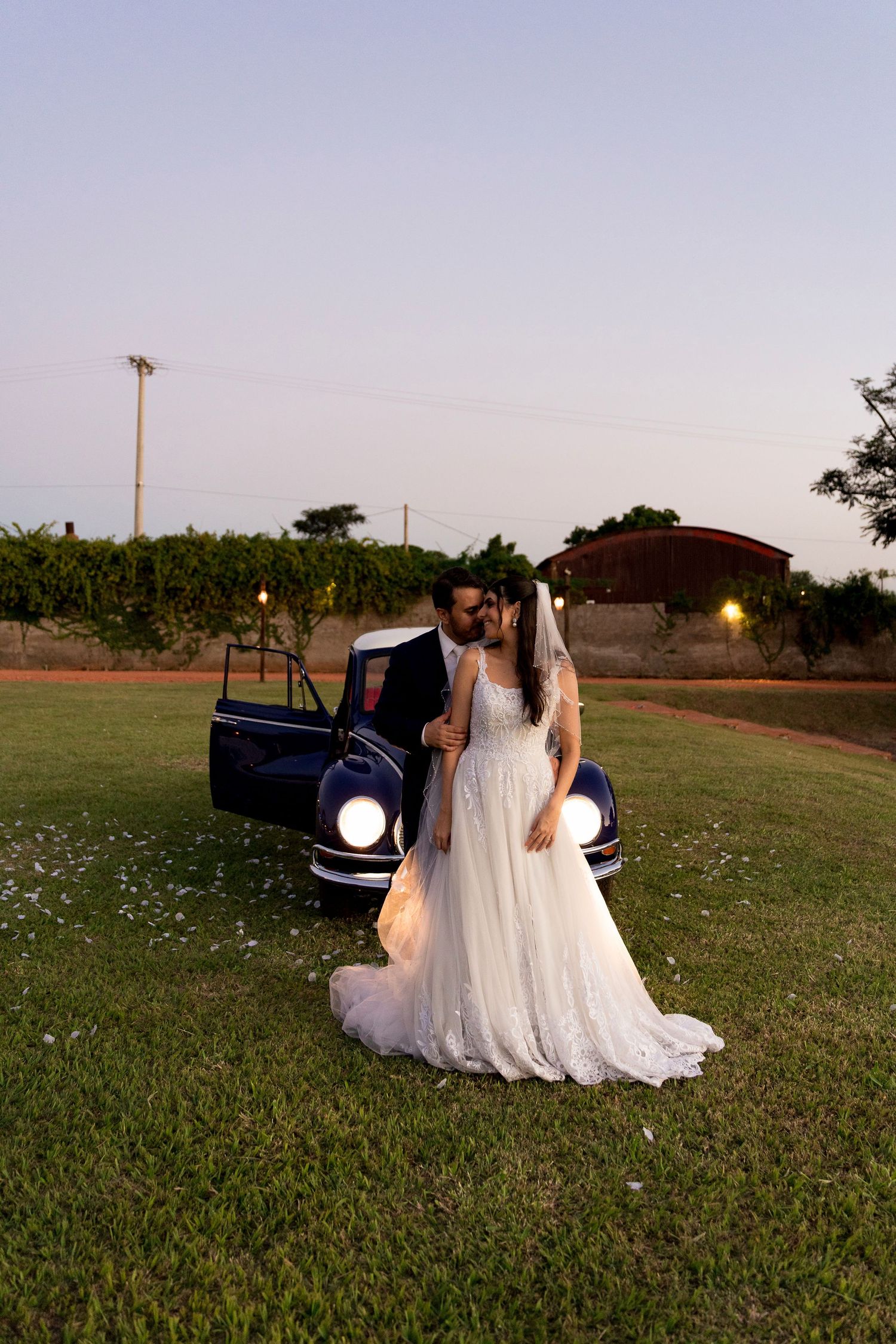 Bride and groom kissing beside a vintage blue car on a green lawn at dusk, with vineyard in the background.