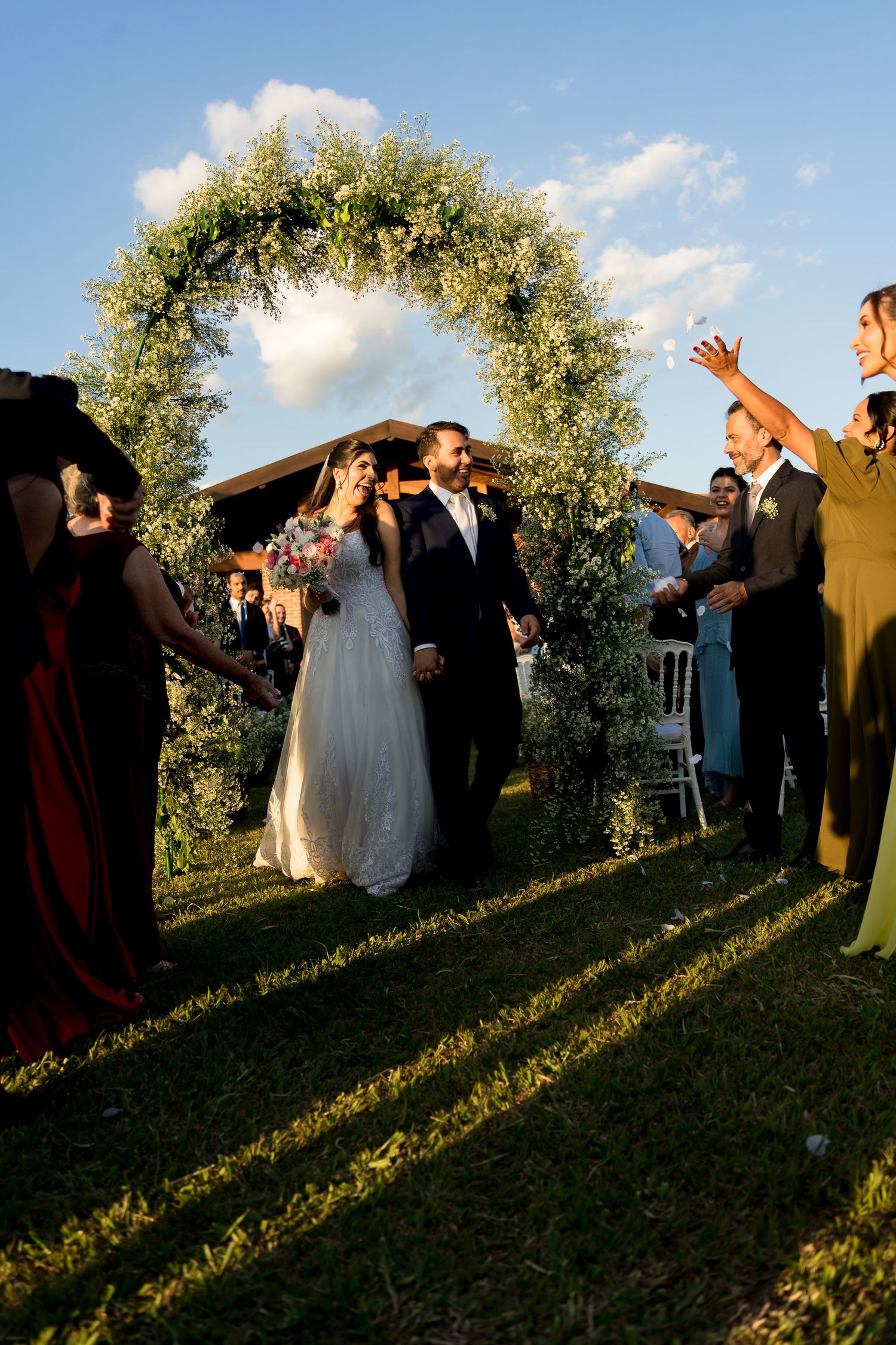 Bride and groom walk through floral arch at outdoor wedding ceremony, guests applauding on sunny day.