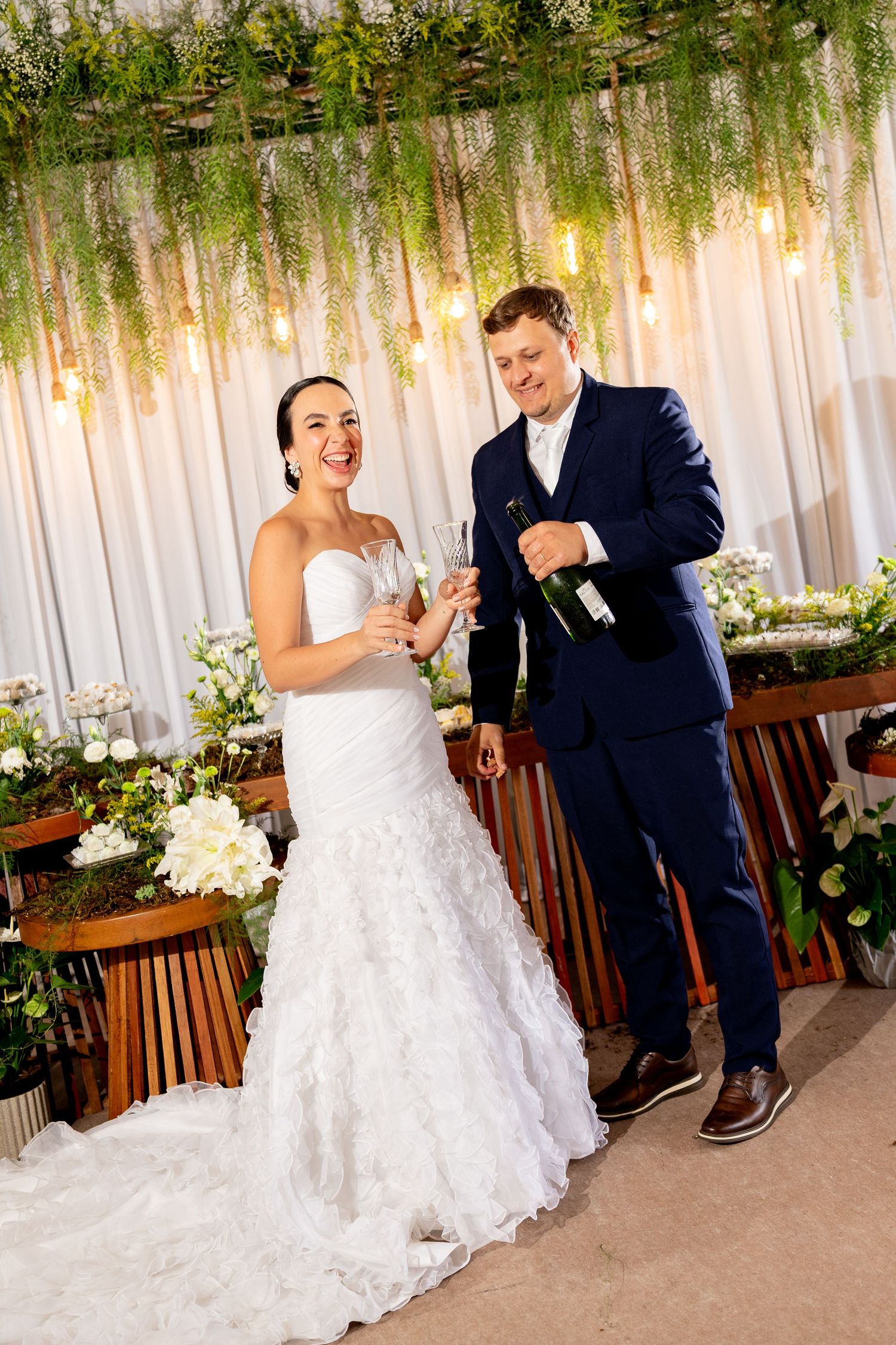 Bride in white gown and groom in navy suit smiling while pouring champagne at elegant floral wedding reception.