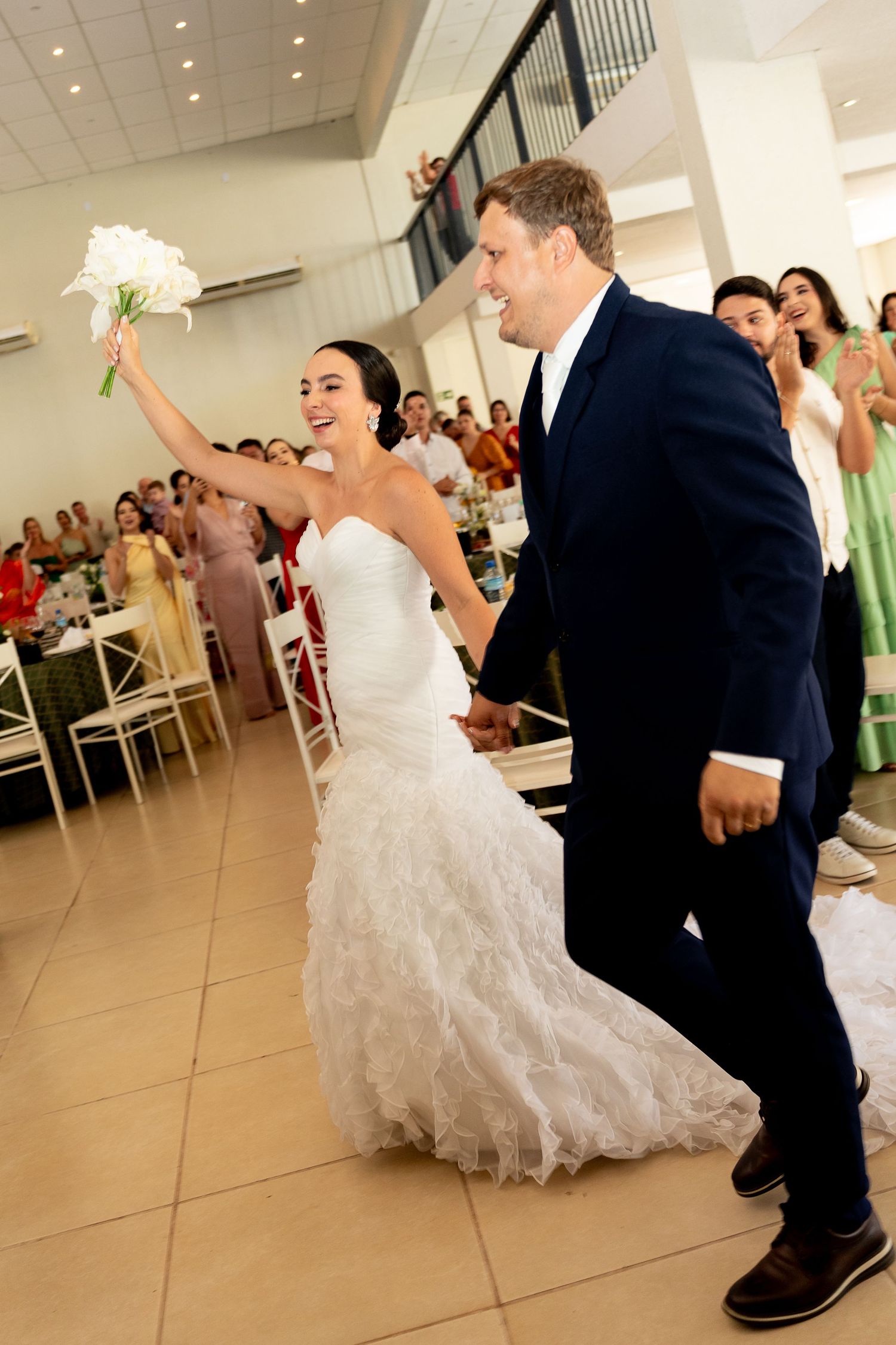 Joyful bride in white ruffled gown raises bouquet while groom in navy suit enters wedding reception hall.