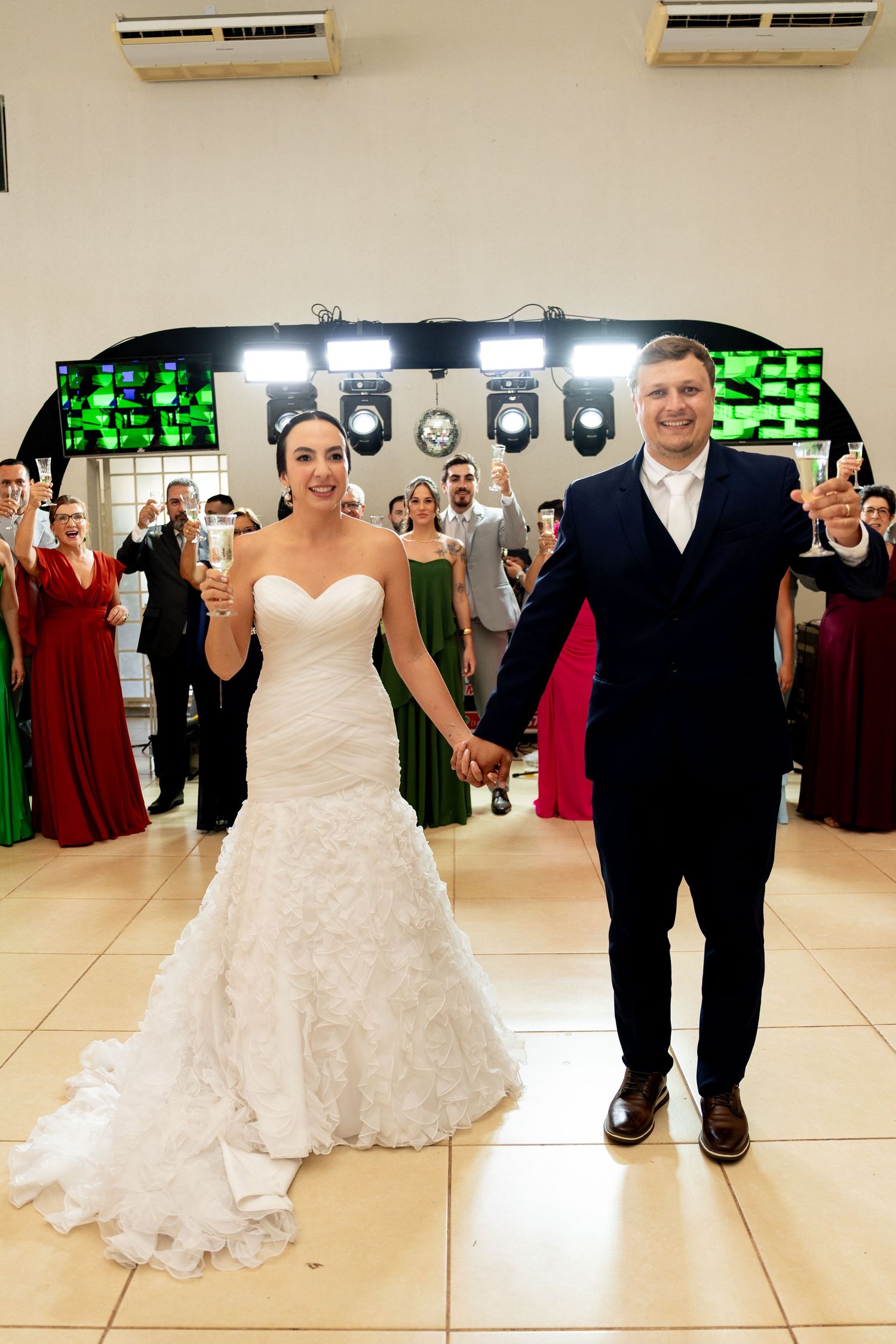 Bride in white gown and groom in navy suit hold hands smiling at wedding reception with guests cheering behind them.