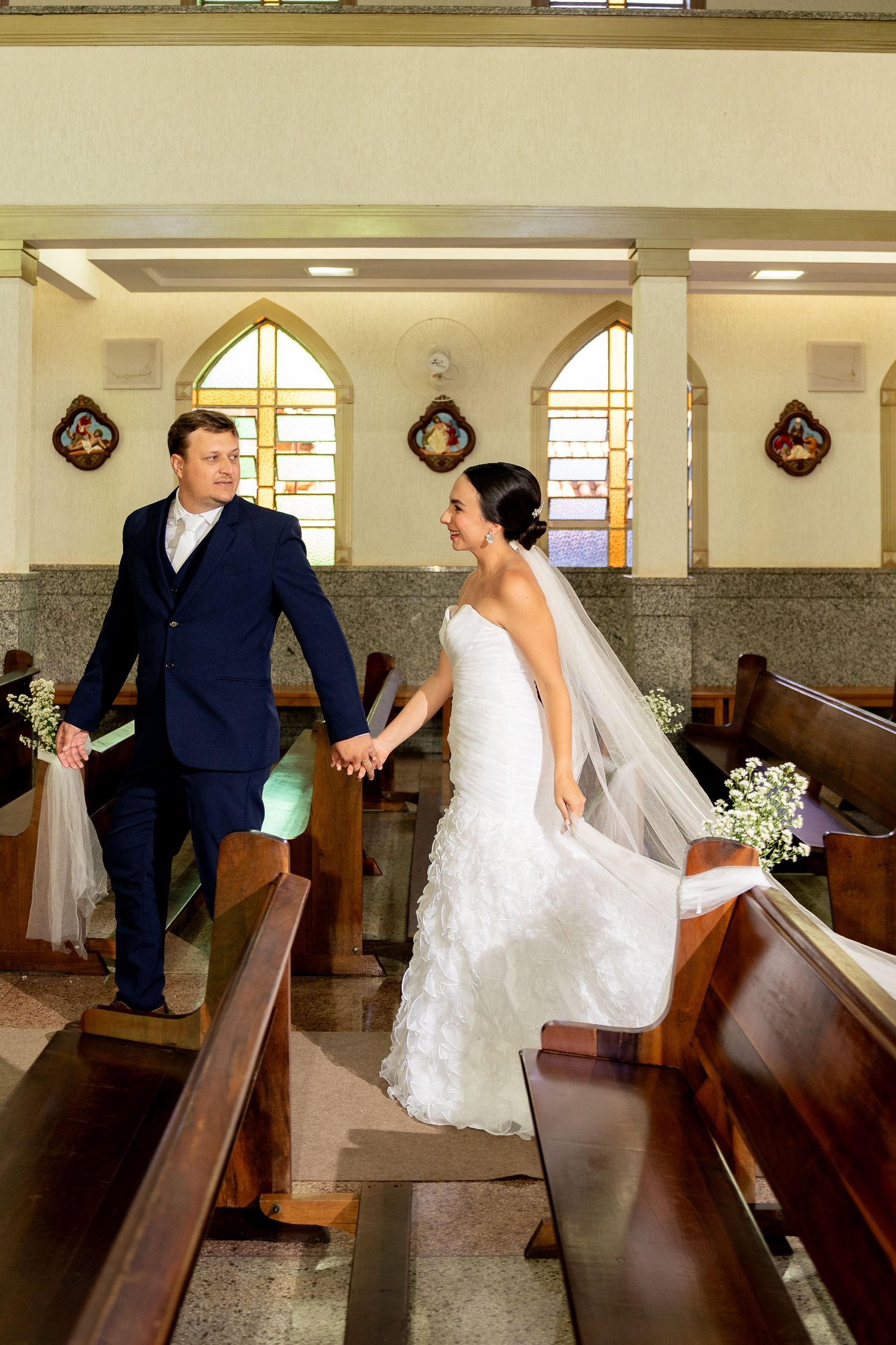 Bride and groom walk hand in hand down church aisle, bride in white gown with veil, groom in navy suit.