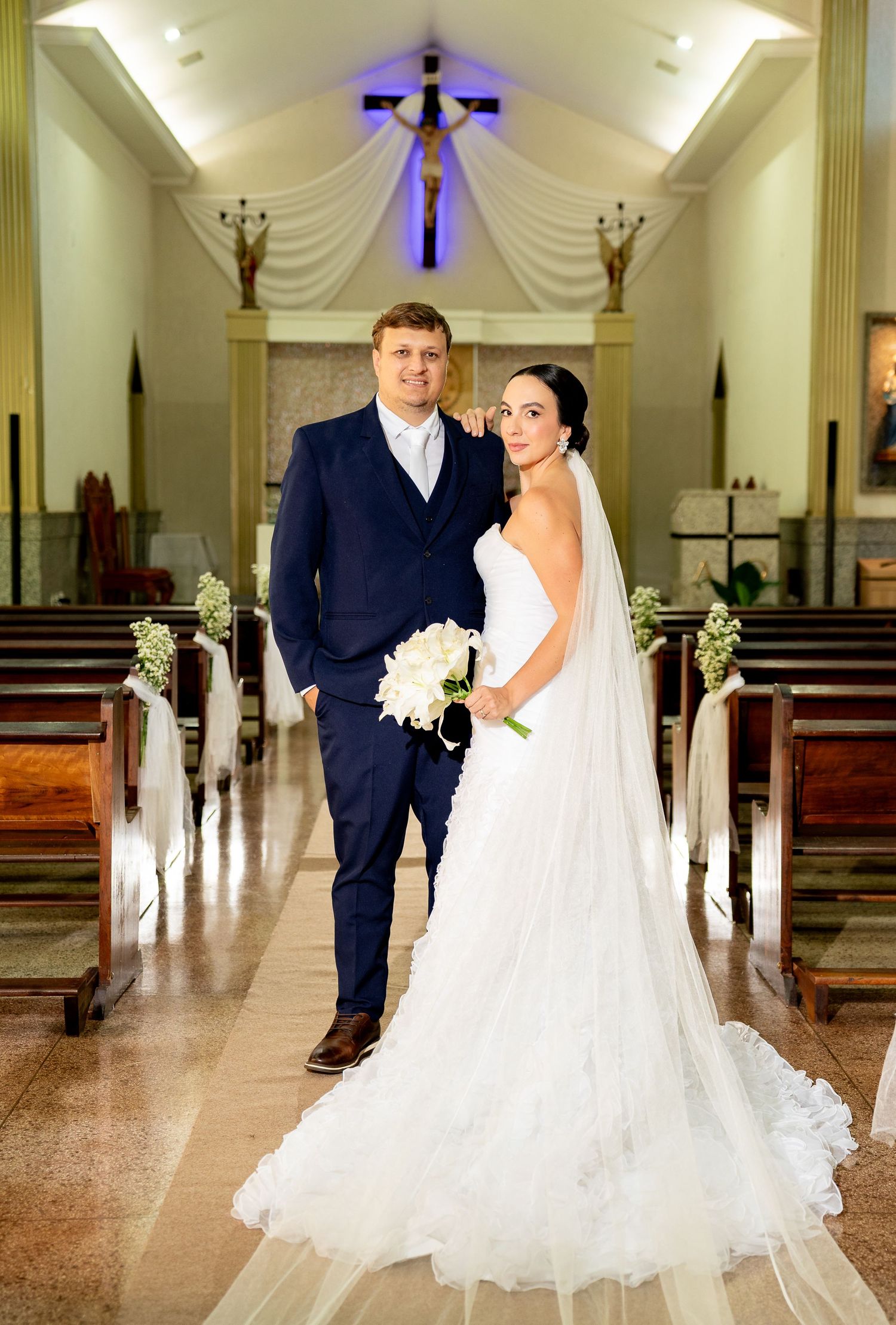 Bride in white gown holding bouquet and groom in navy suit pose together in elegant church aisle after wedding ceremony.