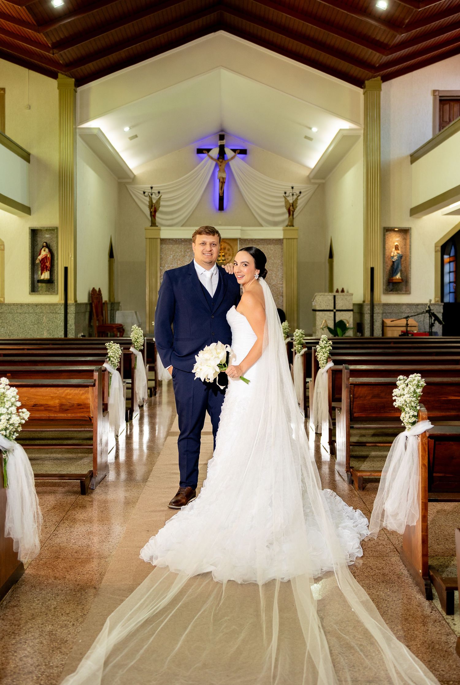 Bride and groom standing together in a beautifully decorated church aisle, bride wearing a white gown with long veil.