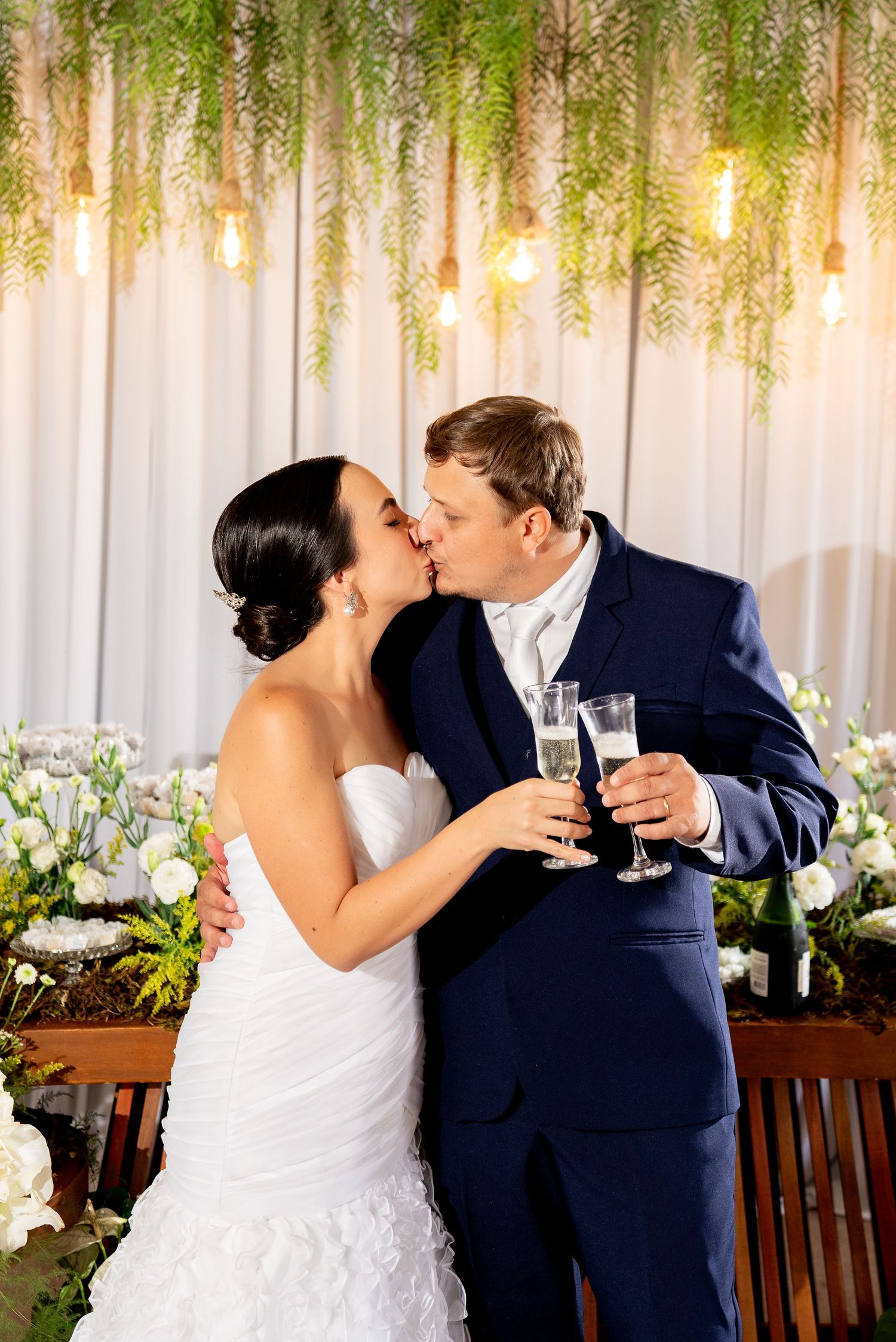 Bride and groom kissing while holding champagne flutes at elegant wedding reception with floral backdrop and hanging lights.