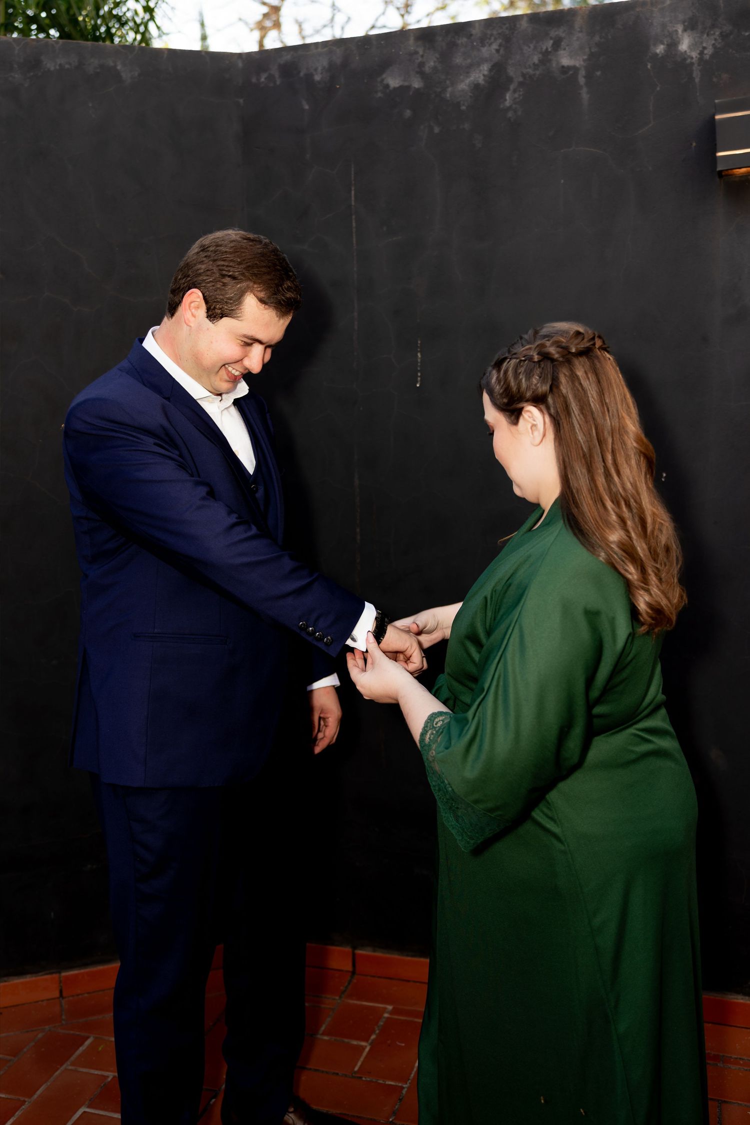 Man in navy suit placing ring on woman in green dress during outdoor proposal against dark wall.