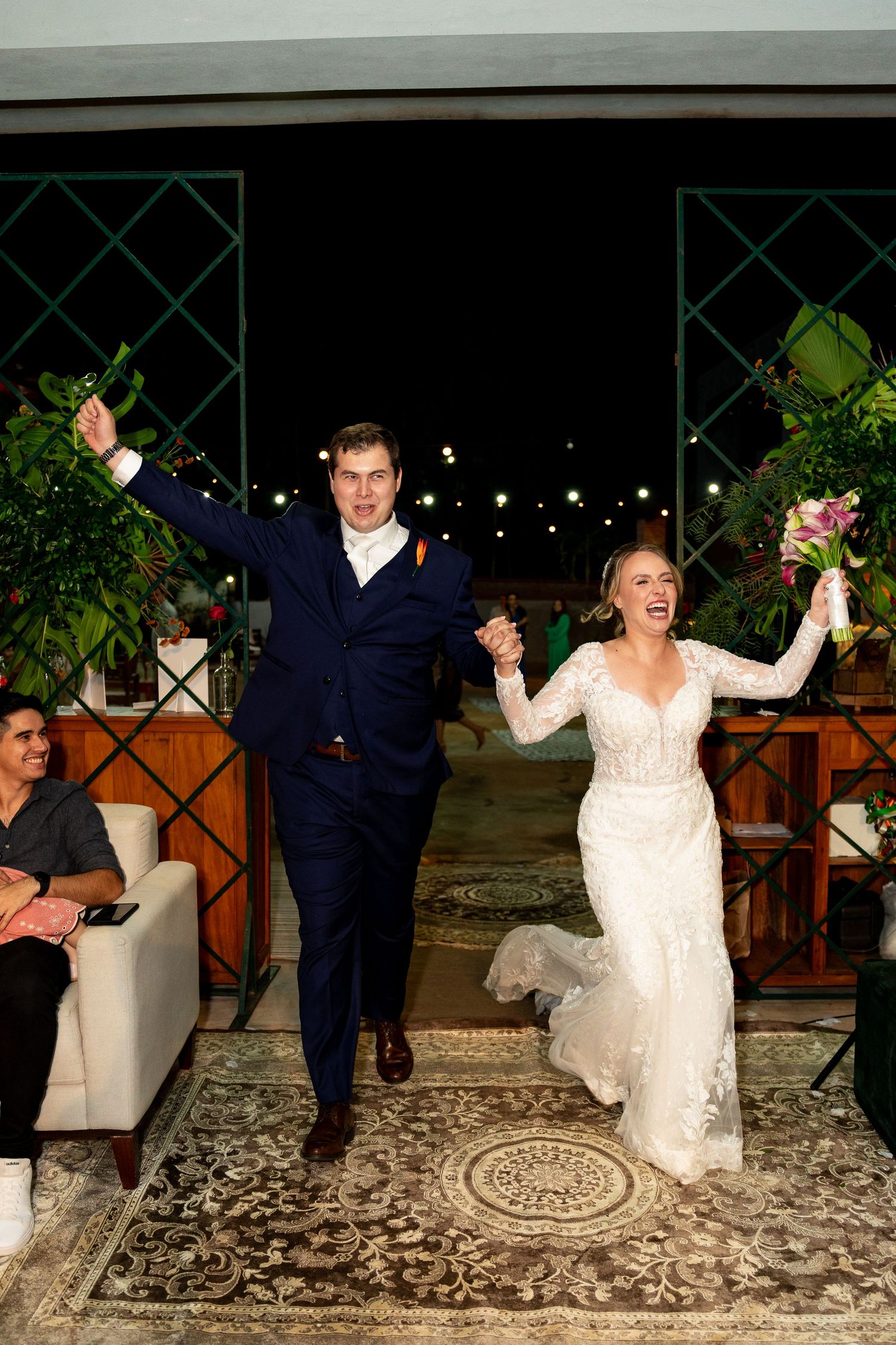 Joyful newlyweds celebrate their grand entrance, bride in lace gown holding bouquet, groom in navy suit, arms raised.