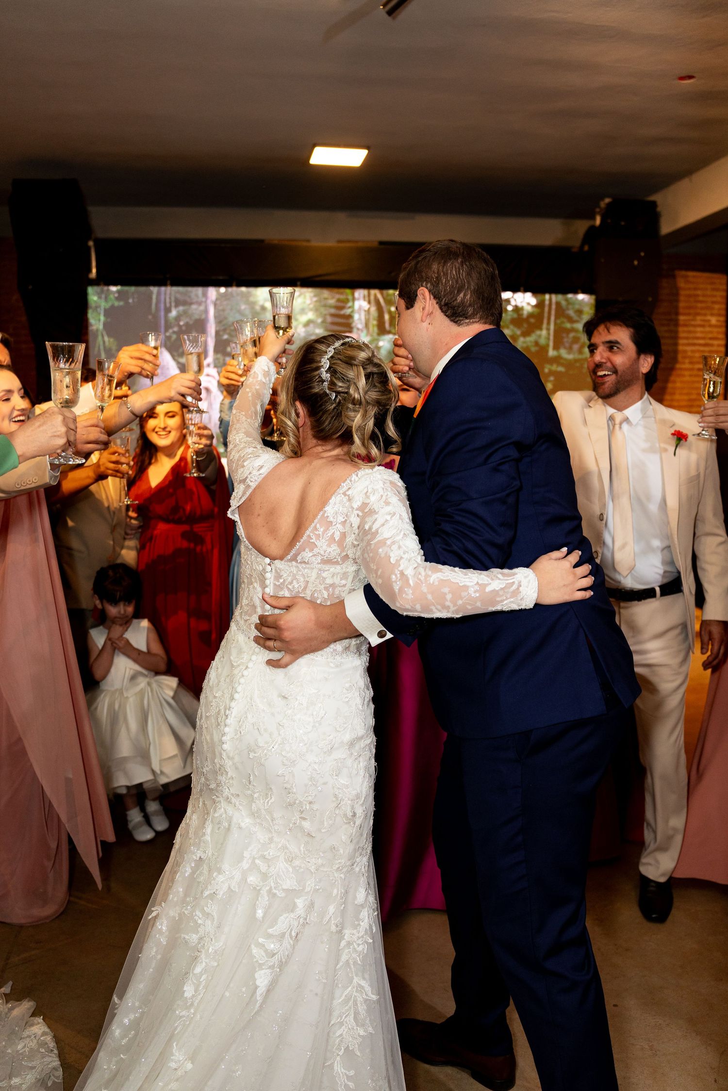 Bride and groom share a kiss surrounded by cheering guests raising champagne glasses at a wedding reception.