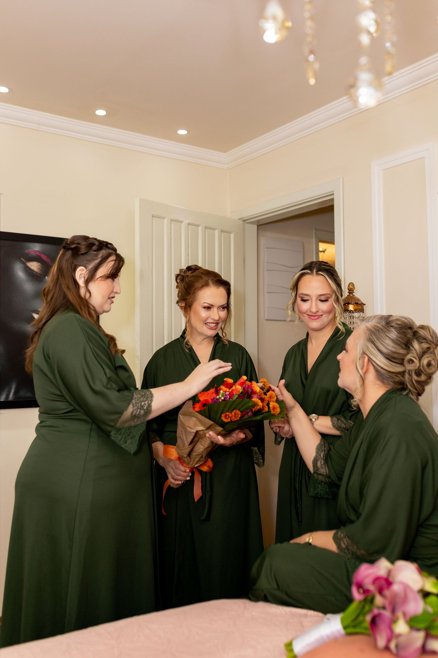 Four bridesmaids in green robes admire a colorful orange and red bouquet while getting ready for a wedding.