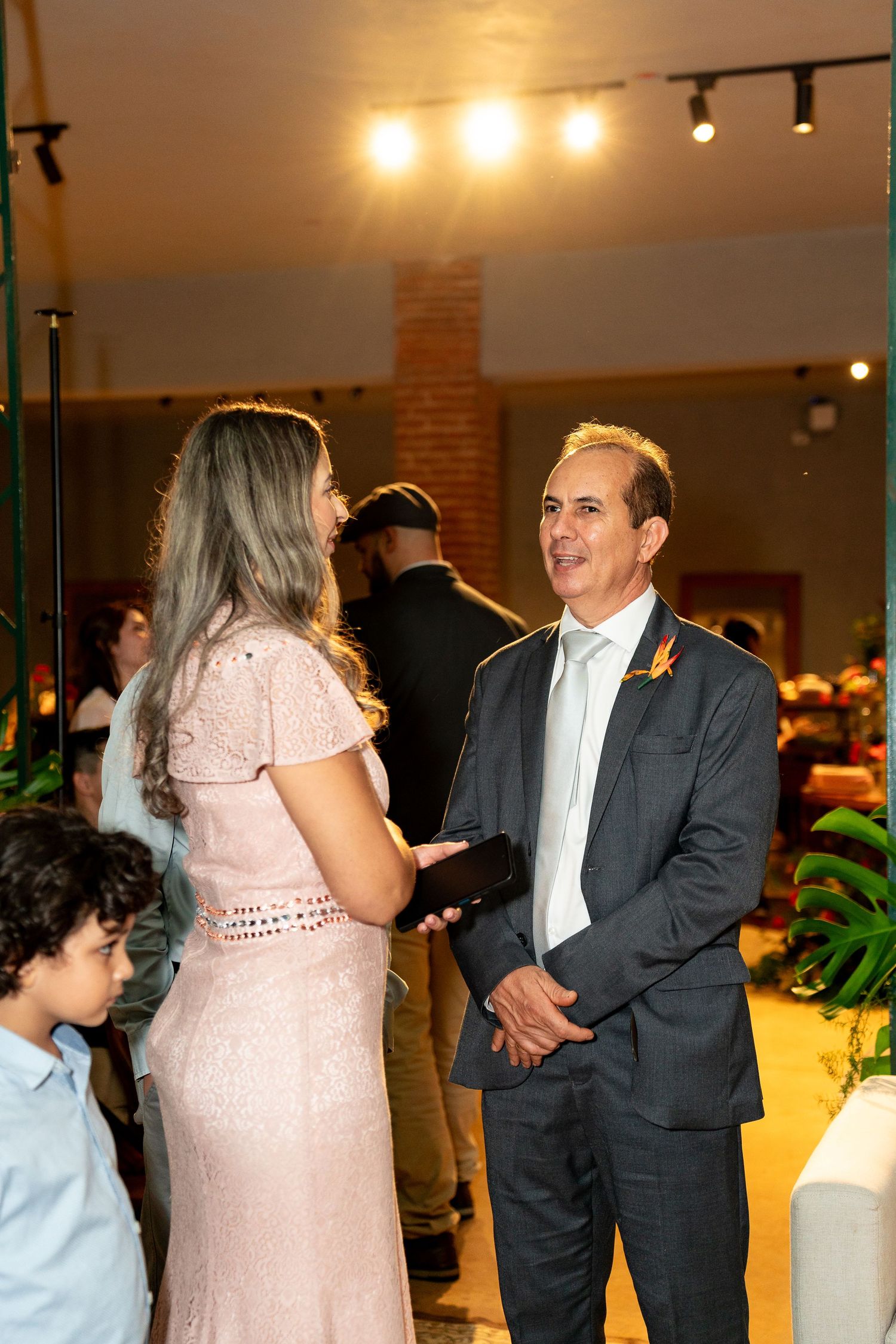A man in a suit and woman in a pink dress converse at an elegant indoor event with warm lighting.