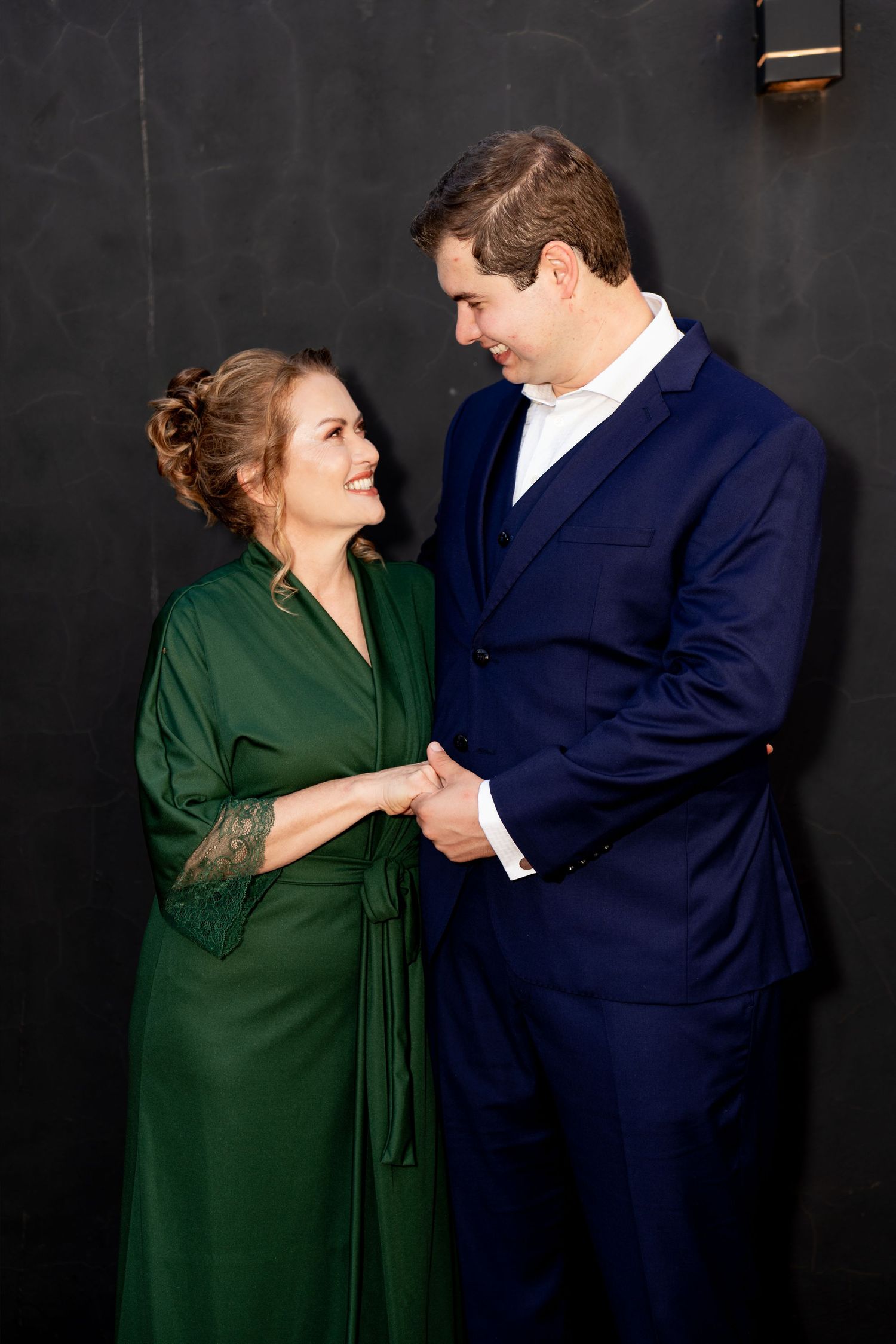 Couple in elegant attire, woman in green dress and man in navy suit, holding hands and smiling at each other.
