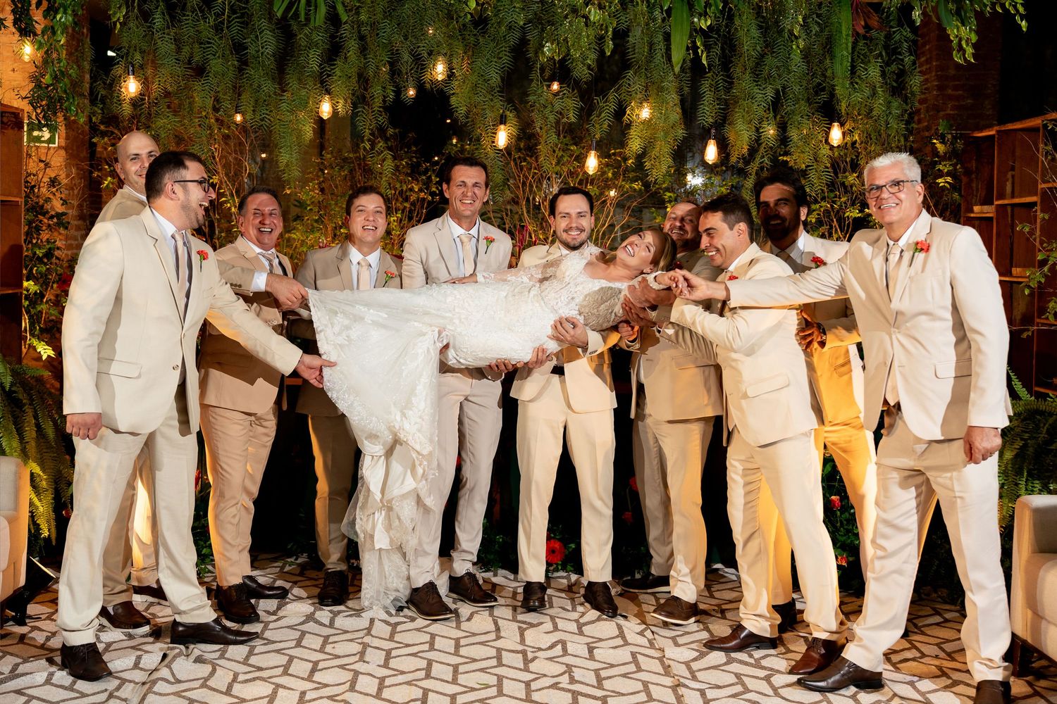 Groomsmen in beige suits lifting the bride at a lush greenery-decorated wedding reception.