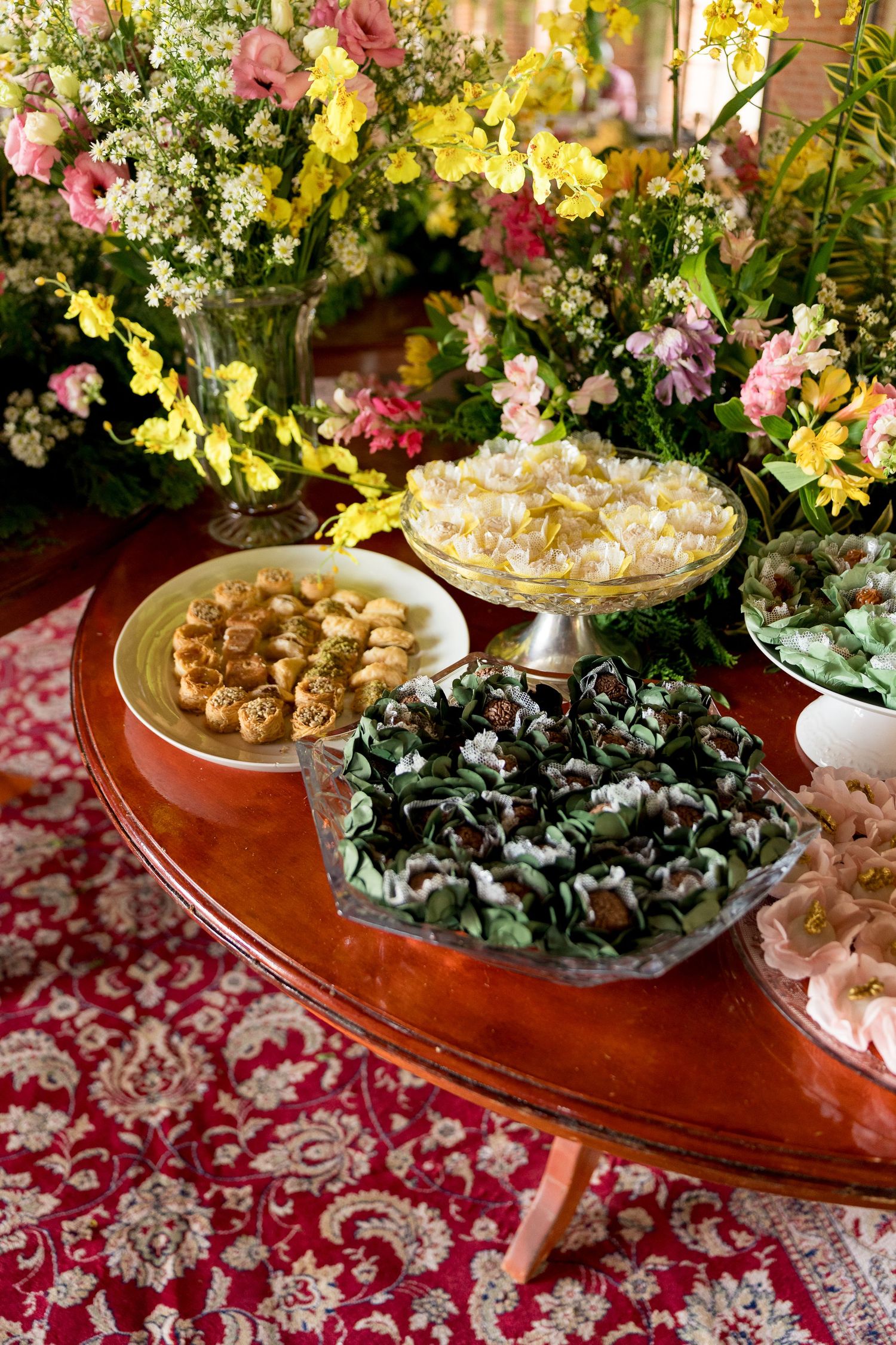 Elegant dessert table with assorted sweets and fresh floral arrangements on a wooden table with red patterned cloth.
