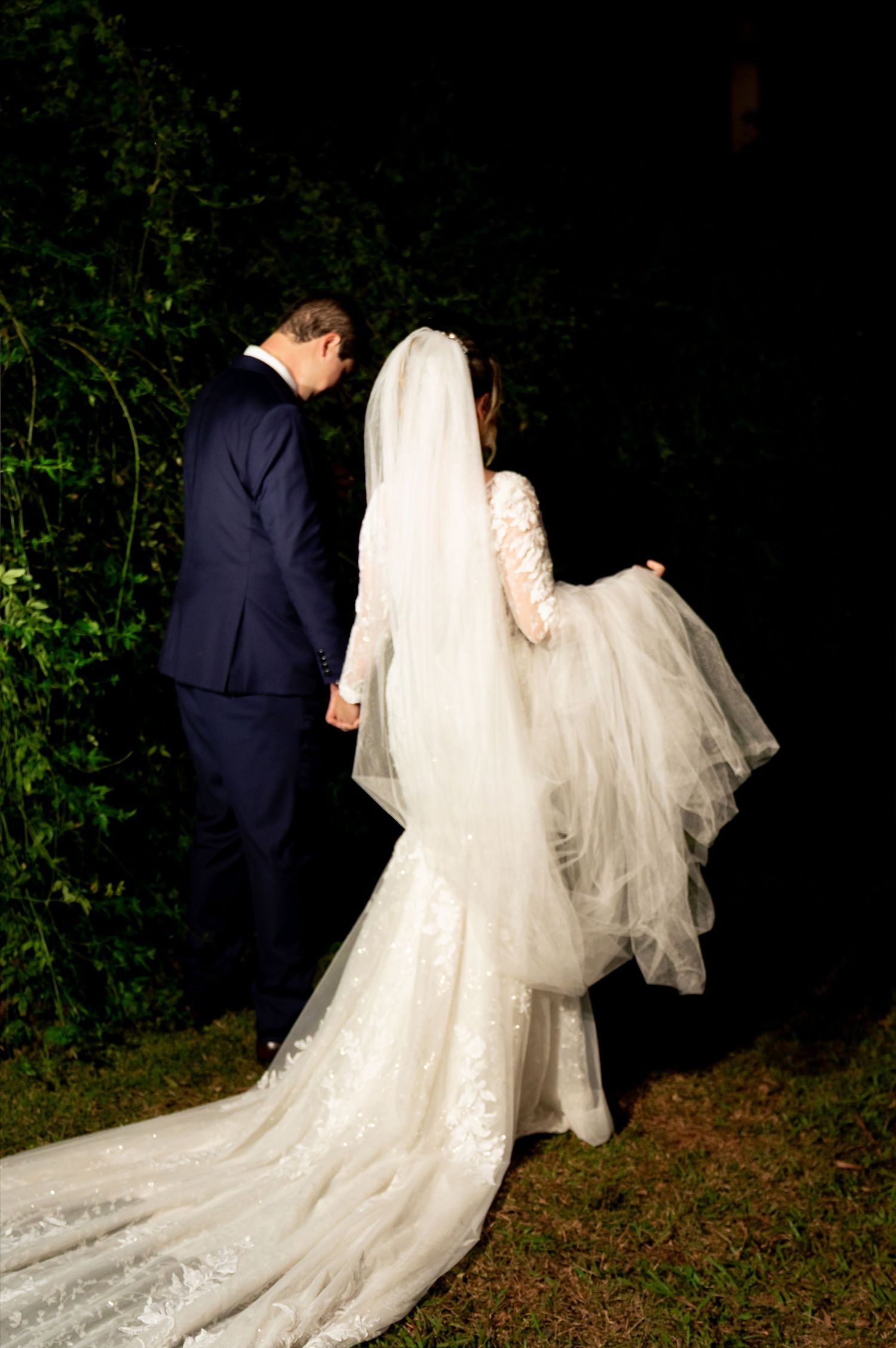 Bride and groom walking hand in hand at night, bride wearing a lace mermaid gown with long veil and train.