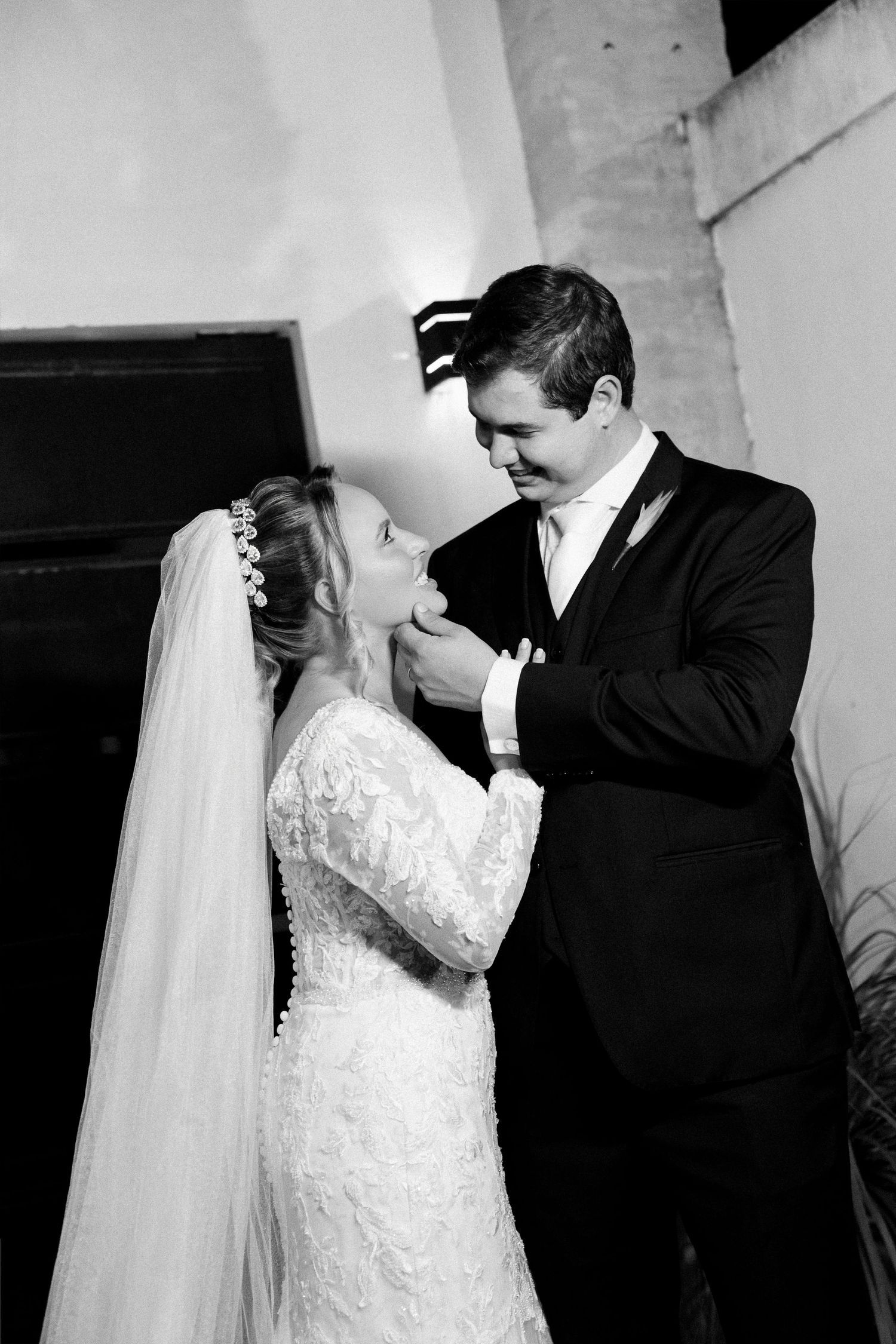 Black and white photo of a bride and groom sharing a tender moment, she in lace gown with veil, he in dark suit.