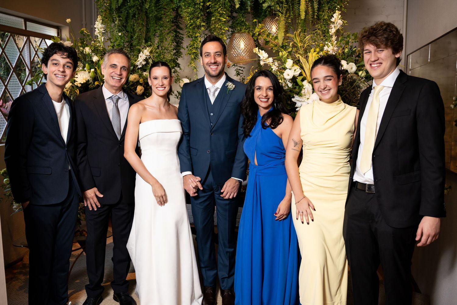 Wedding party of seven smiling guests posing together indoors with floral greenery backdrop.