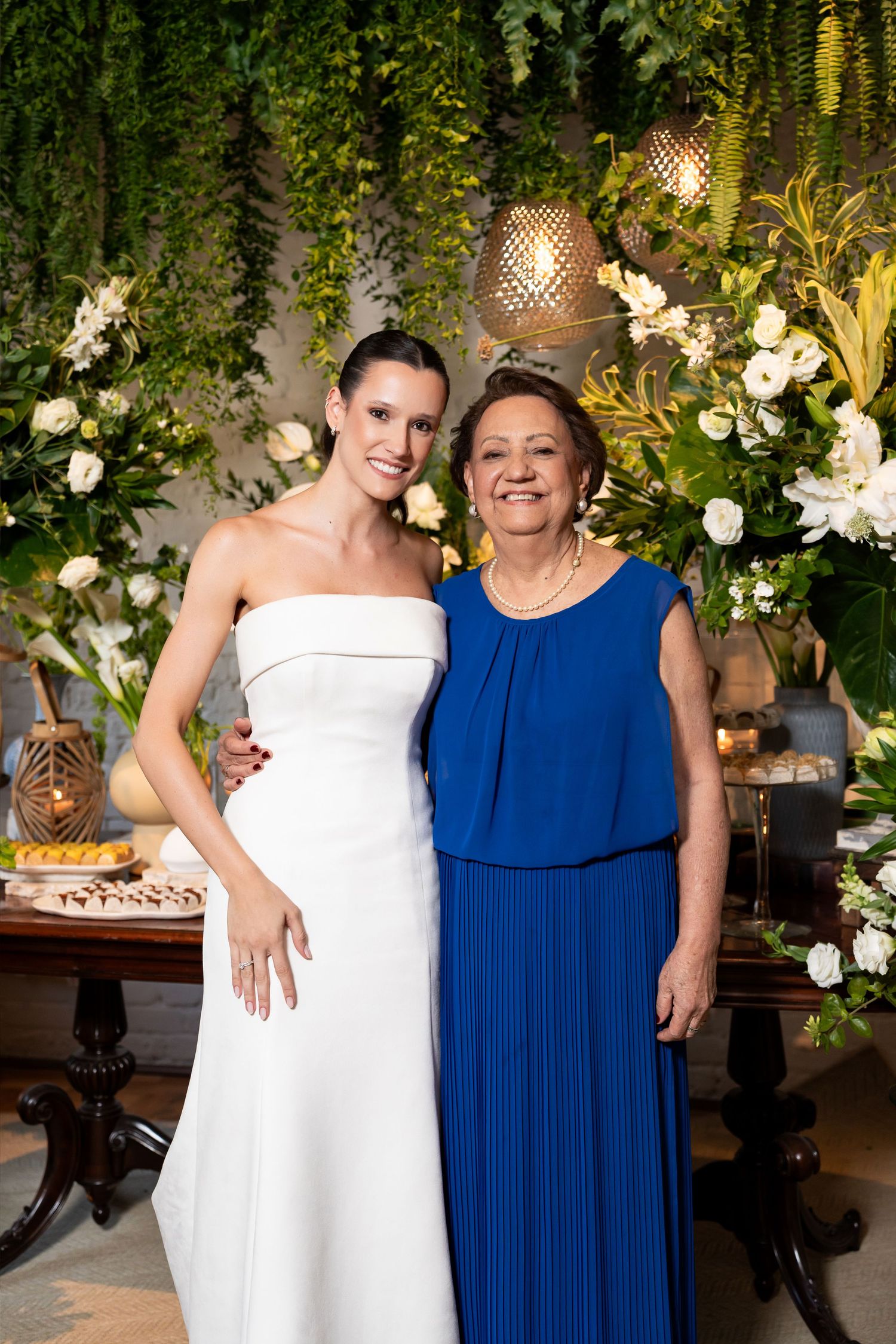 Bride in white strapless gown poses with older woman in royal blue dress at floral wedding reception.