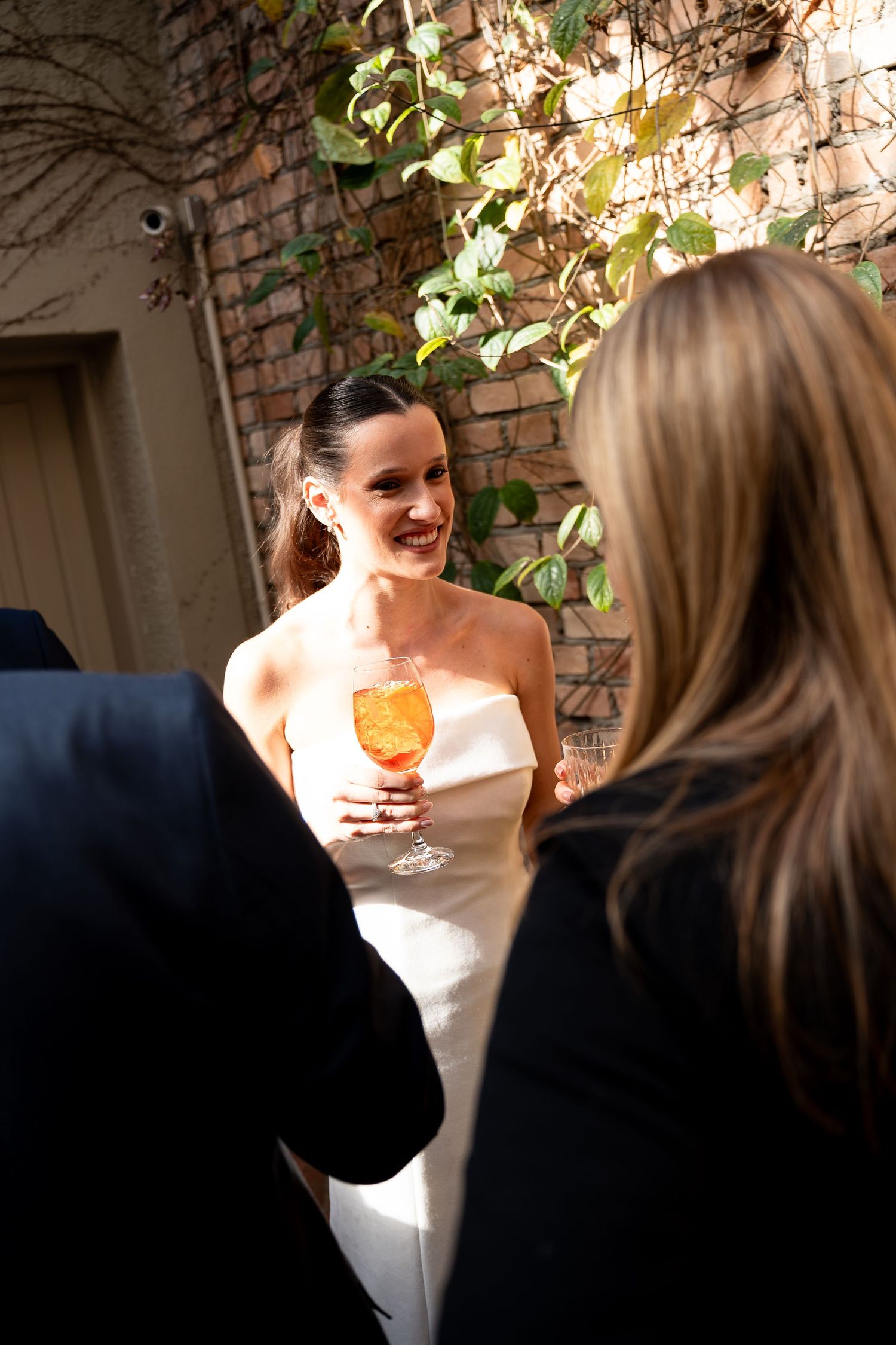Smiling bride in white strapless gown holding an Aperol spritz cocktail, chatting with guests at an outdoor wedding reception.