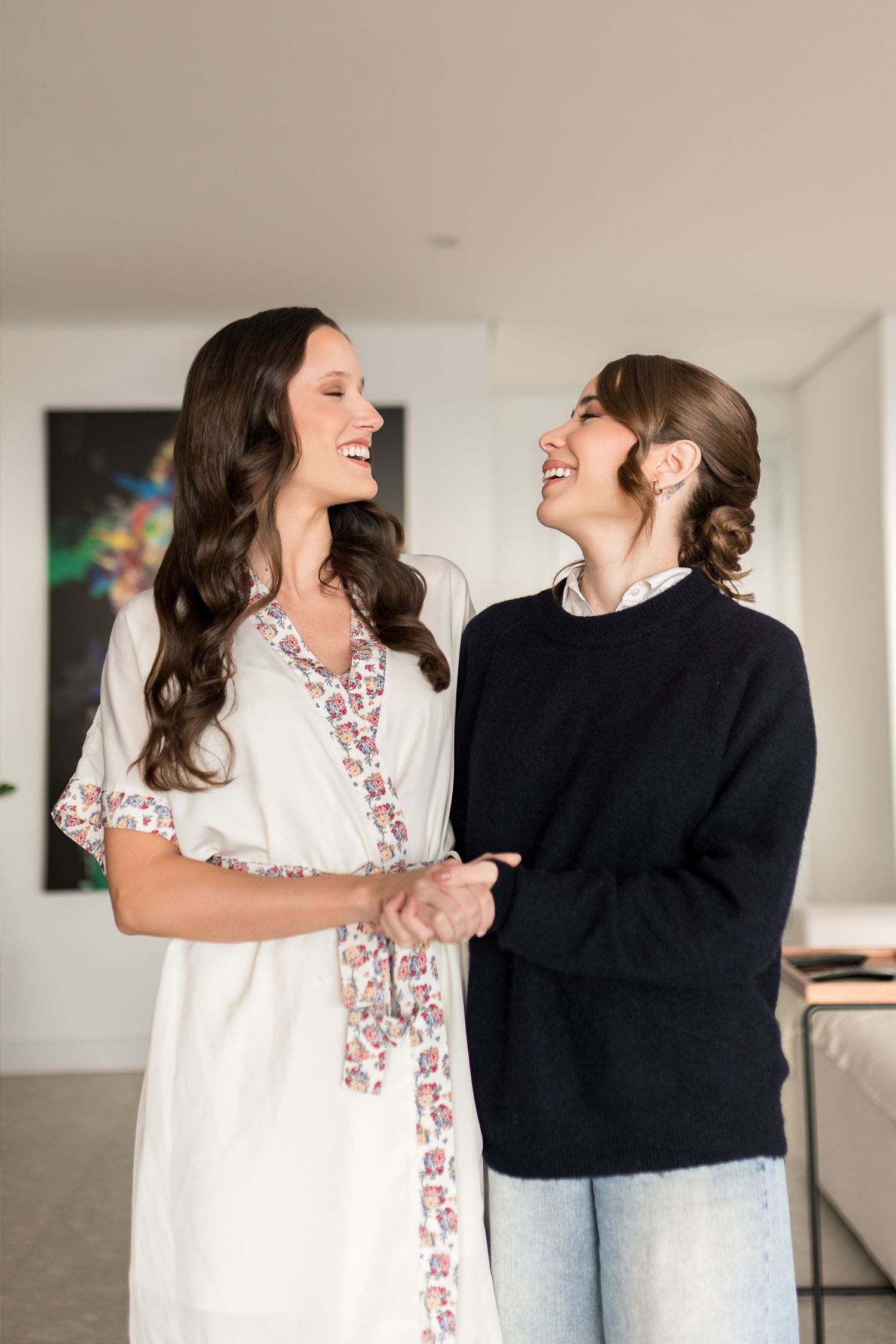 Two women laughing and holding hands indoors, one in a floral white dress and one in a navy sweater.