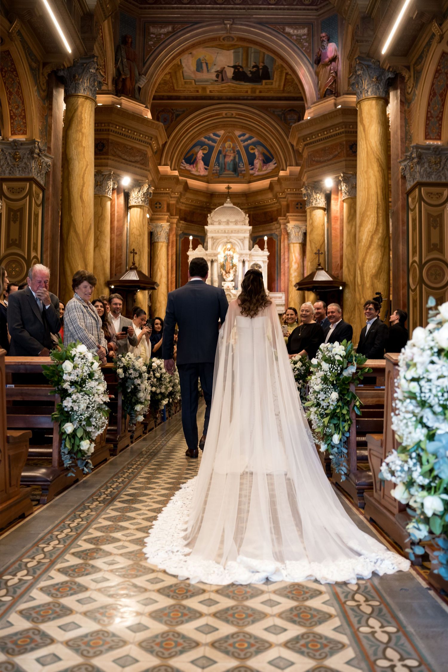 Bride and groom walk down ornate church aisle adorned with white floral arrangements, bride wearing elegant cape wedding gown.