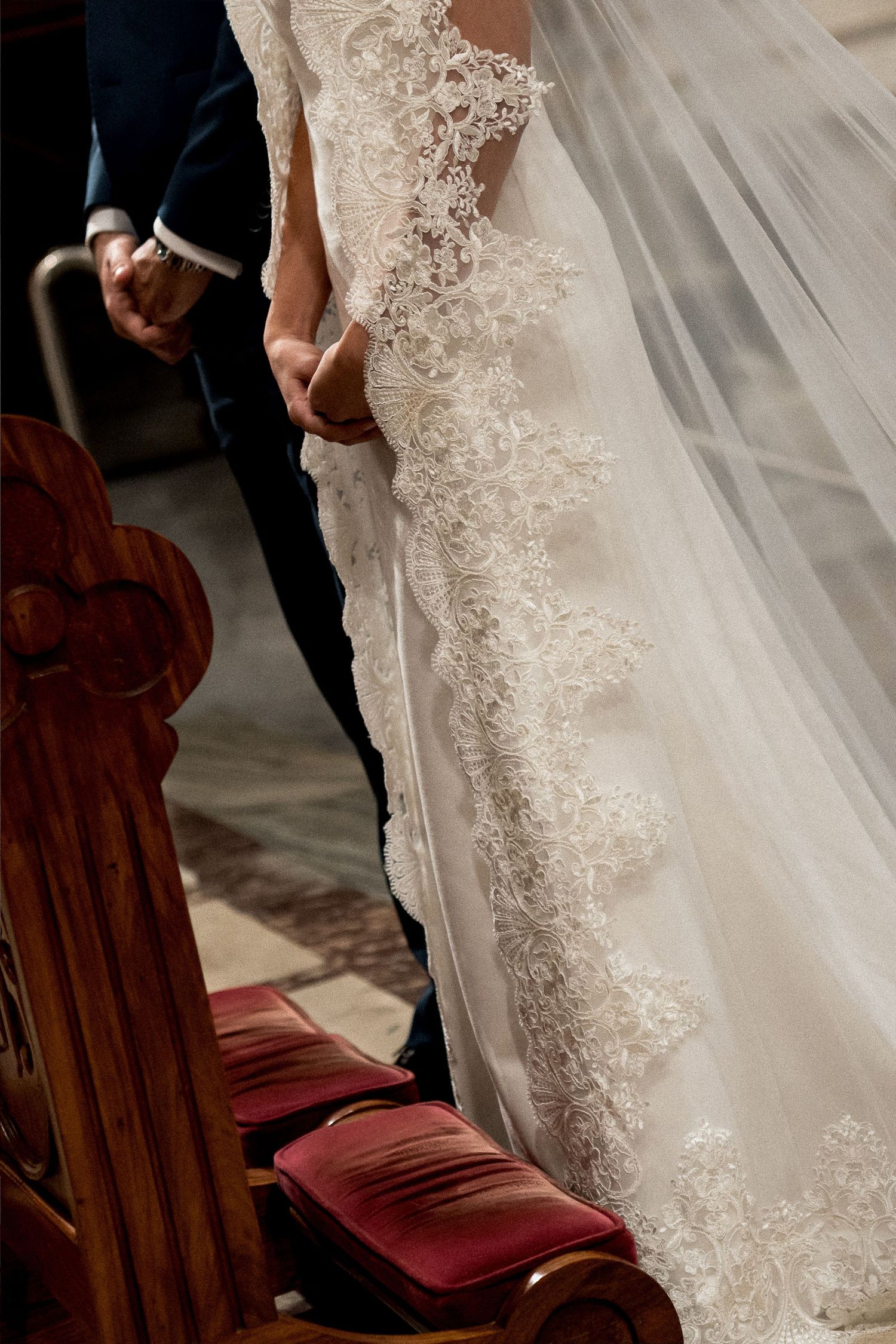 Bride in lace wedding gown and veil holding groom's hand beside ornate wooden church pew with red cushion.