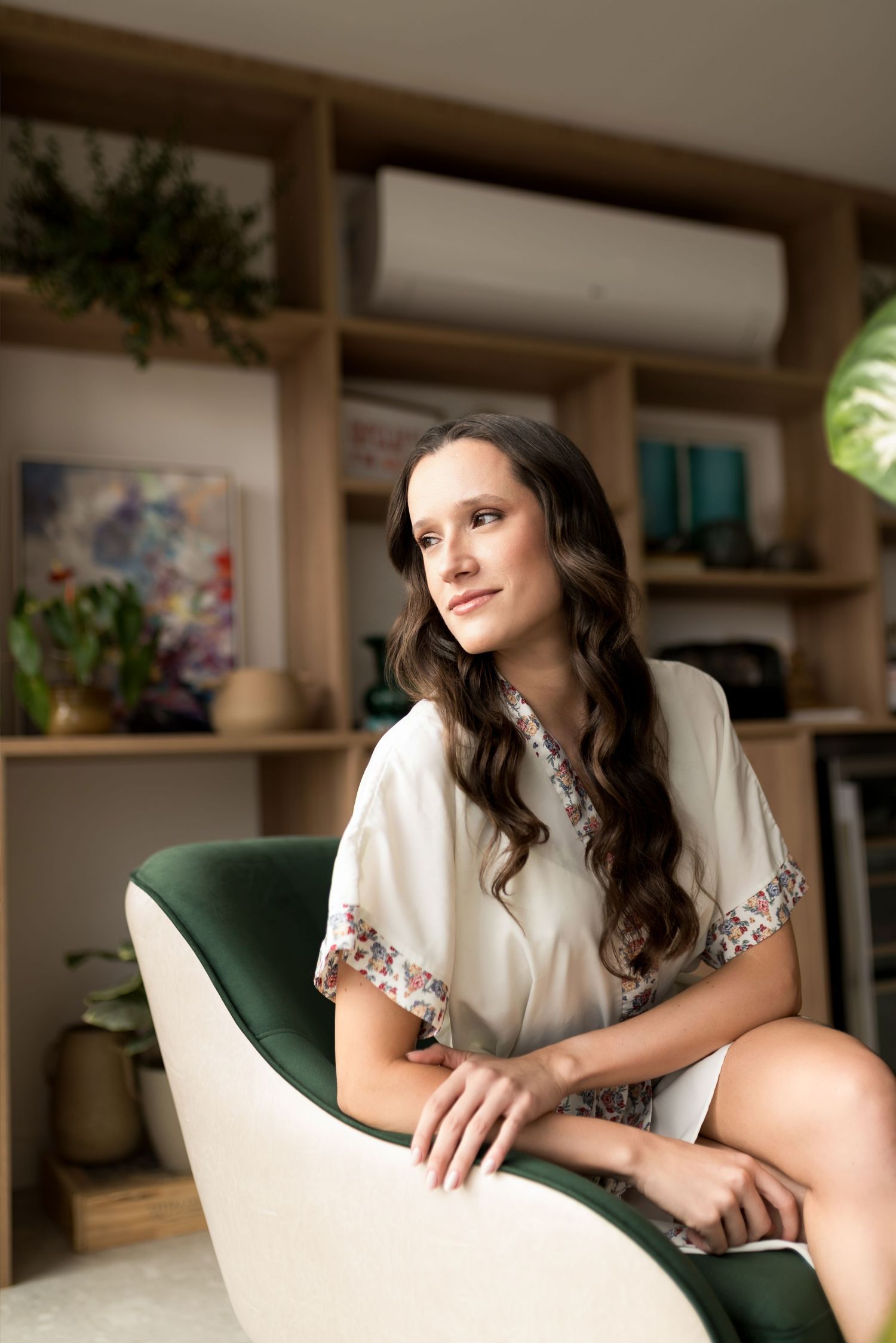 Woman in white blouse sitting in green chair, smiling thoughtfully in a stylish room with shelves and plants.