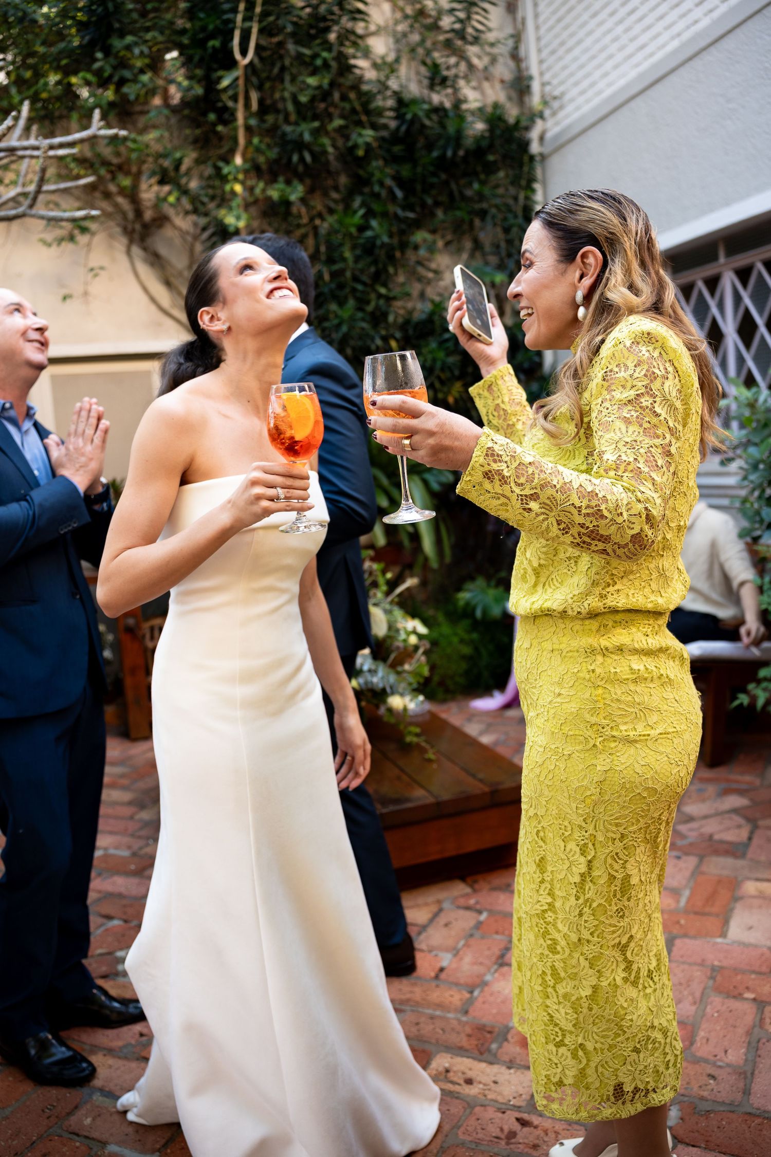 Bride in white gown and guest in yellow lace dress toasting with wine glasses at an outdoor wedding reception.