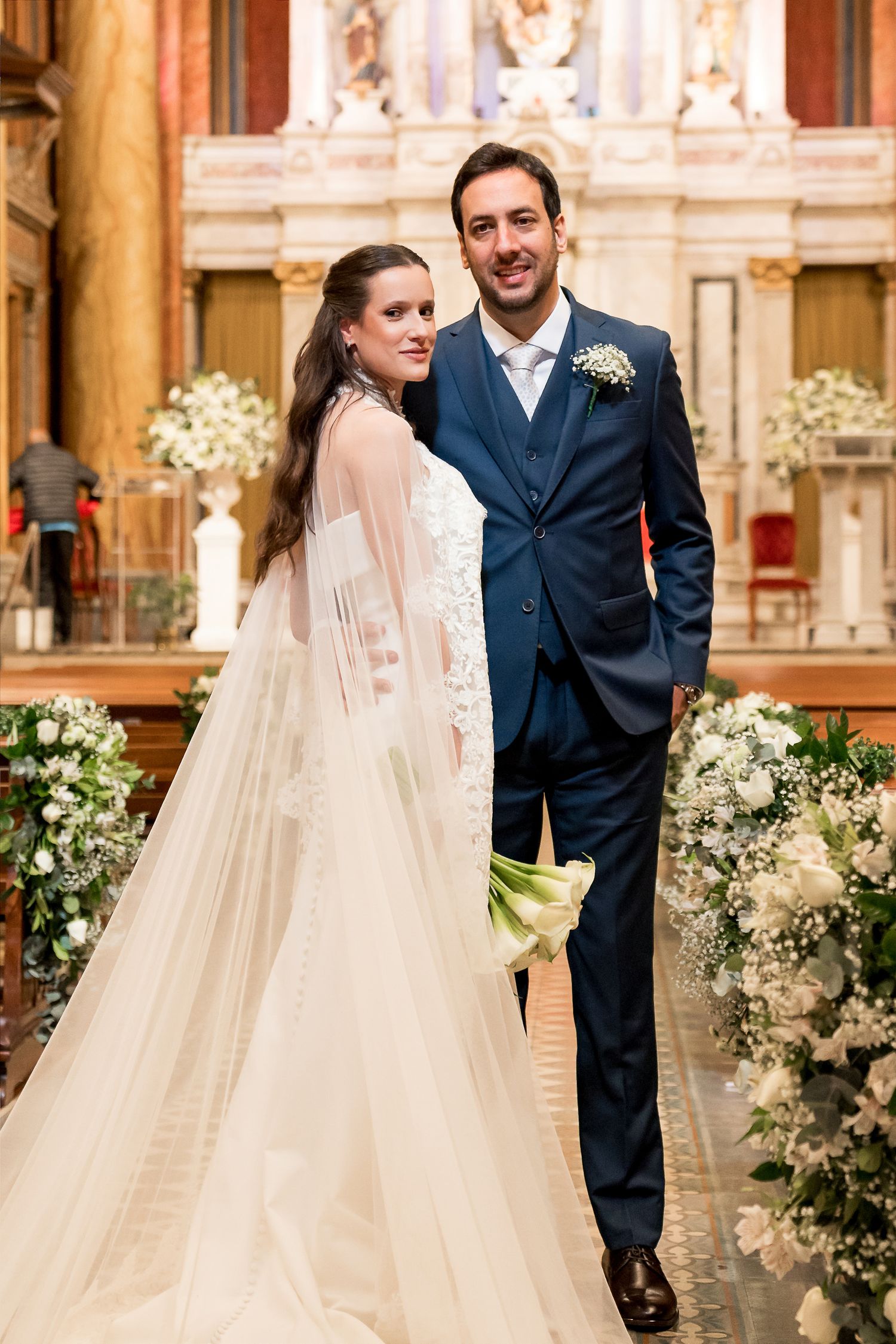 Bride in white lace gown and groom in navy suit pose together at elegant church wedding ceremony.