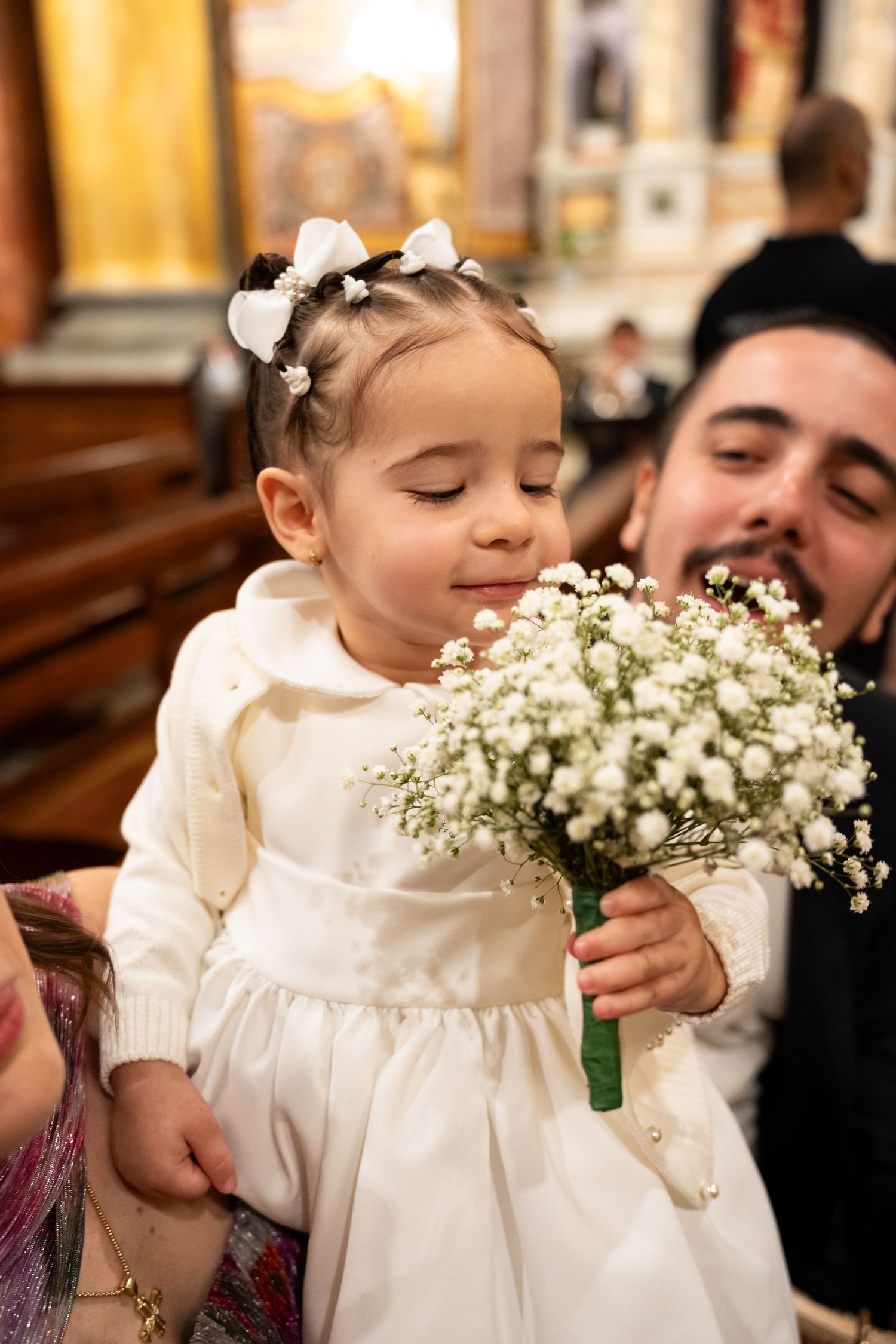 Young flower girl in white dress holds baby's breath bouquet at a wedding ceremony in a church.