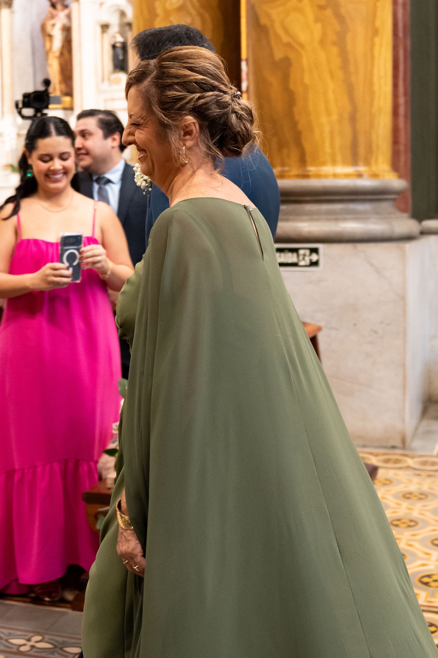 Woman in elegant olive green cape gown smiling at a formal event inside an ornate building.