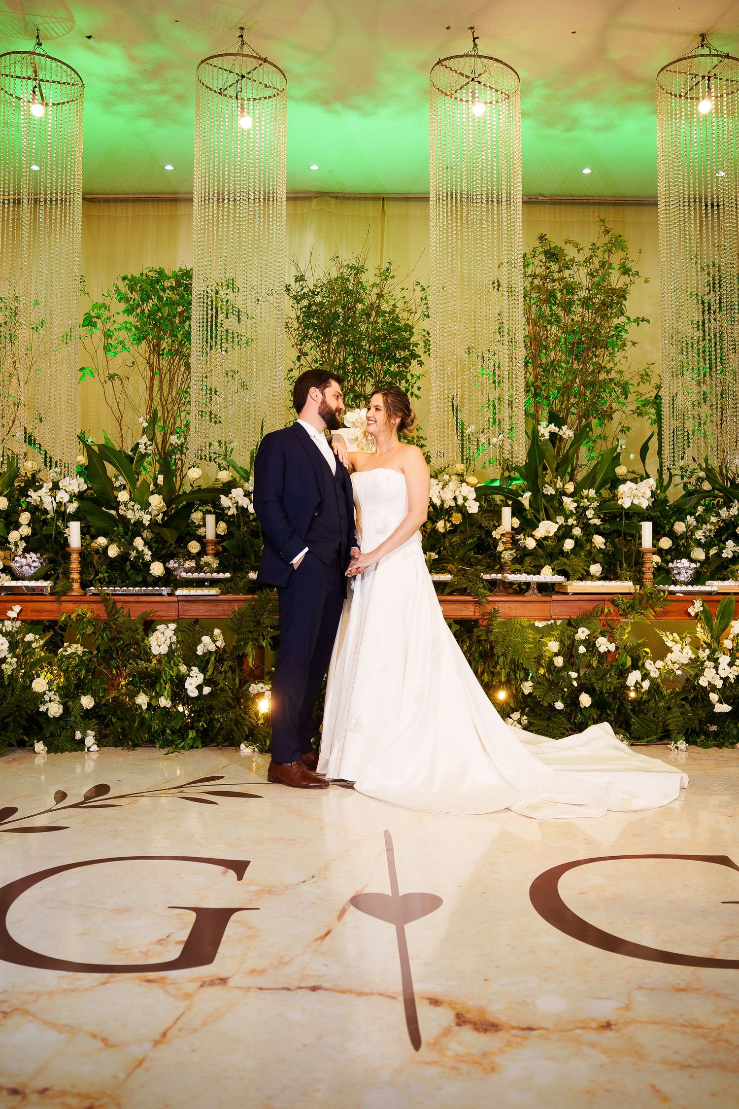 Bride and groom share a romantic kiss at elegant wedding altar with lush greenery, white florals, and crystal chandeliers.