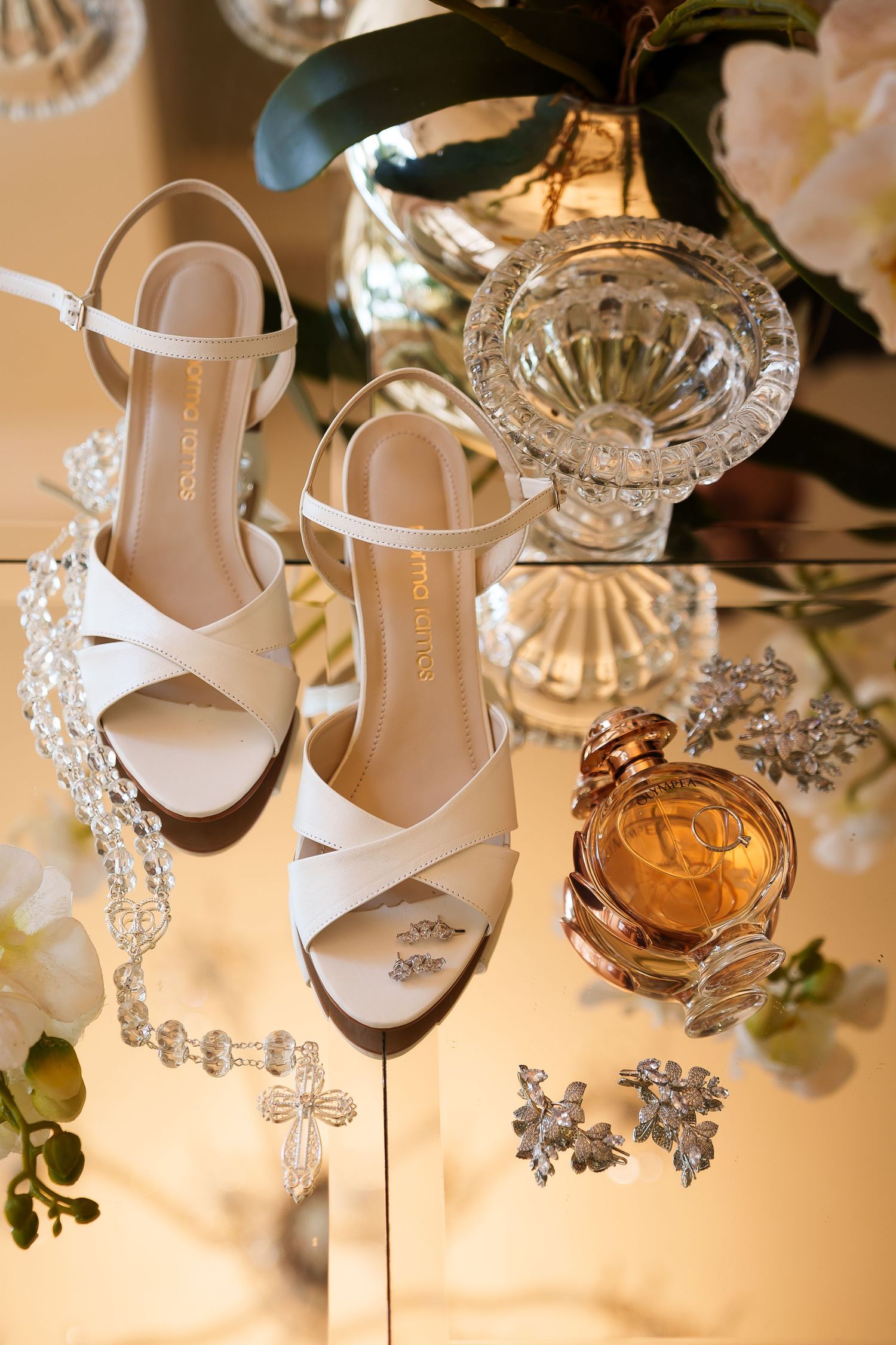 White bridal heels with crystal jewelry, floral earrings, and amber perfume bottle arranged on a mirrored surface with flowers.